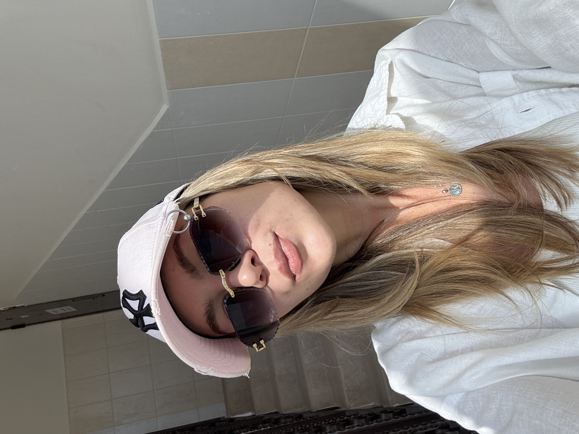 An individual, facing the camera, poses for a selfie outdoors. The person wears a pink baseball cap with a black logo, sunglasses, and a white collared shirt. A necklace with a blue pendant is visible. The background consists of outdoor stairs and tiled walls. The scene appears to be in a residential area.