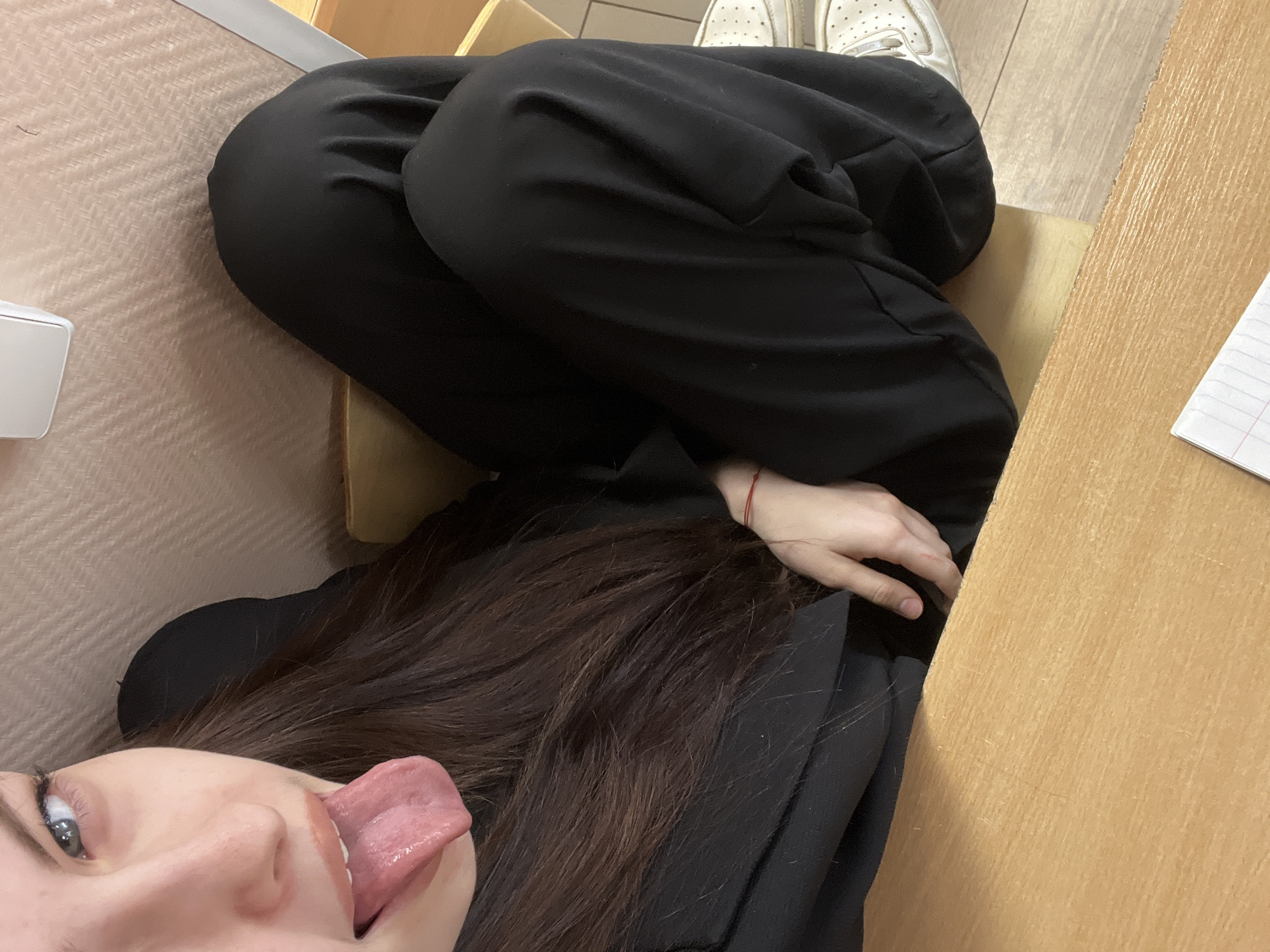 A close-up shot captures a person sticking their tongue out toward the camera, presumably in an indoor setting. Their dark hair cascades beside their face. The person is wearing loose black pants. A portion of their hand is visible near the right side of the frame. The person is sitting in a wooden chair, which is positioned near a light-colored wooden desk or table. A glimpse of white sneakers can be seen in the upper part of the picture. A piece of lined paper is situated on the wooden surface. The location is unknown and cannot be determined from the picture.