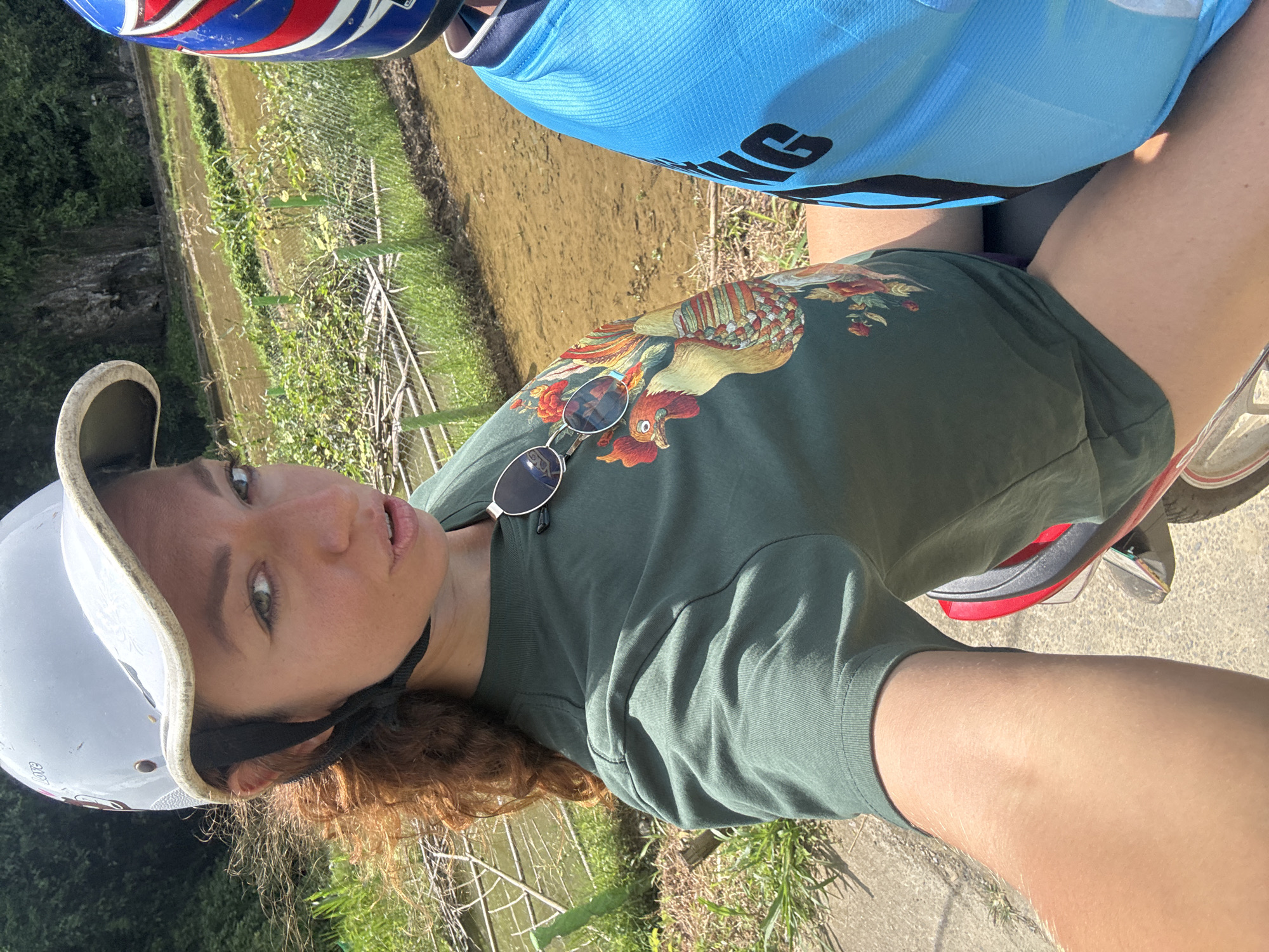 The image shows a woman with curly red hair, wearing a white helmet and a green t-shirt with a colorful rooster design. She is also wearing sunglasses. She is looking at the camera with a neutral expression. The woman appears to be sitting on a bicycle or motorcycle, with part of the vehicle visible in the lower right corner. She is outdoors, with a flooded rice paddy and greenery in the background. The sun is shining, suggesting it is daytime. The scene is likely set in a rural area, given the presence of rice paddies. The woman is positioned in a way that suggests she is taking a selfie. Her blue cycling jersey is also visible, indicating she may be on a cycling trip. The location context provided is Pù Luông Commune, Vietnam, which aligns with the rural, rice paddy landscape.