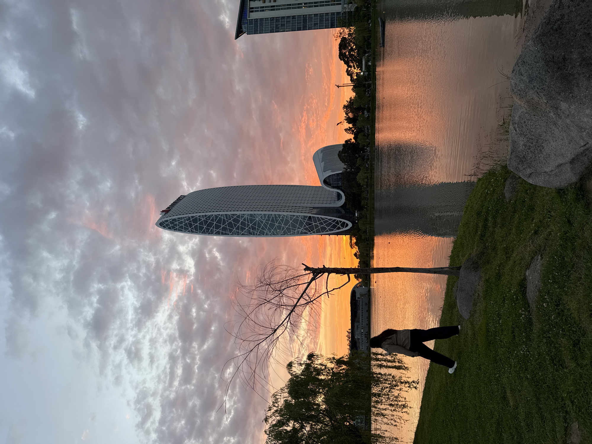 The image shows a lakeside view in Batumi, Georgia, at sunset. A modern, oval-shaped building with a lattice-like design is positioned on the shore. The sky is filled with colorful clouds, reflecting orange and pink hues onto the water's surface. A lone tree with bare branches stands near the edge, and a person is standing on the grassy bank. The building's design and the overall setting suggest a contemporary urban landscape.