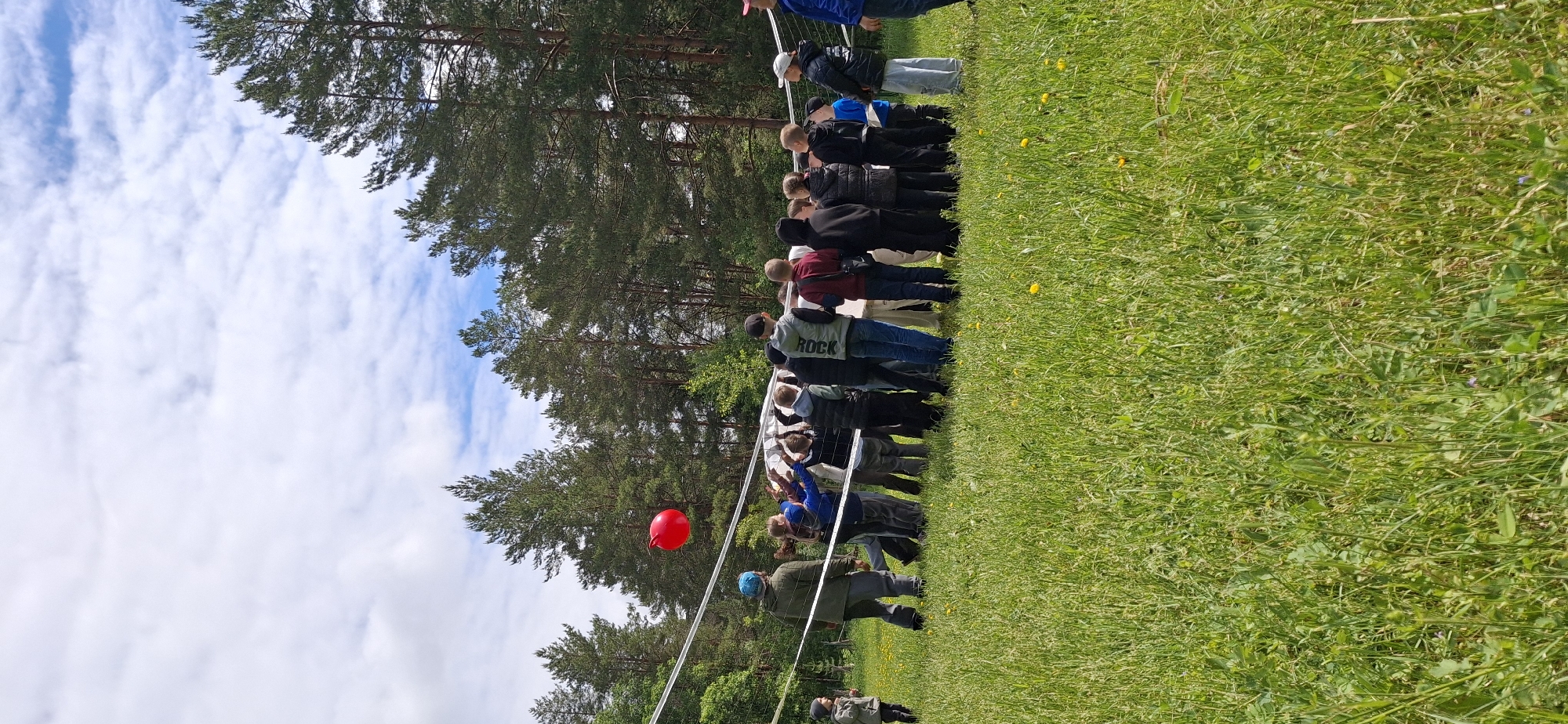 The image shows a group of people gathered in a grassy area in Прикамский, Russia. The group is standing behind a white net or fence, possibly observing an event taking place in the adjacent field. A red balloon is visible near the net. The setting includes a backdrop of trees under a sky with scattered clouds.