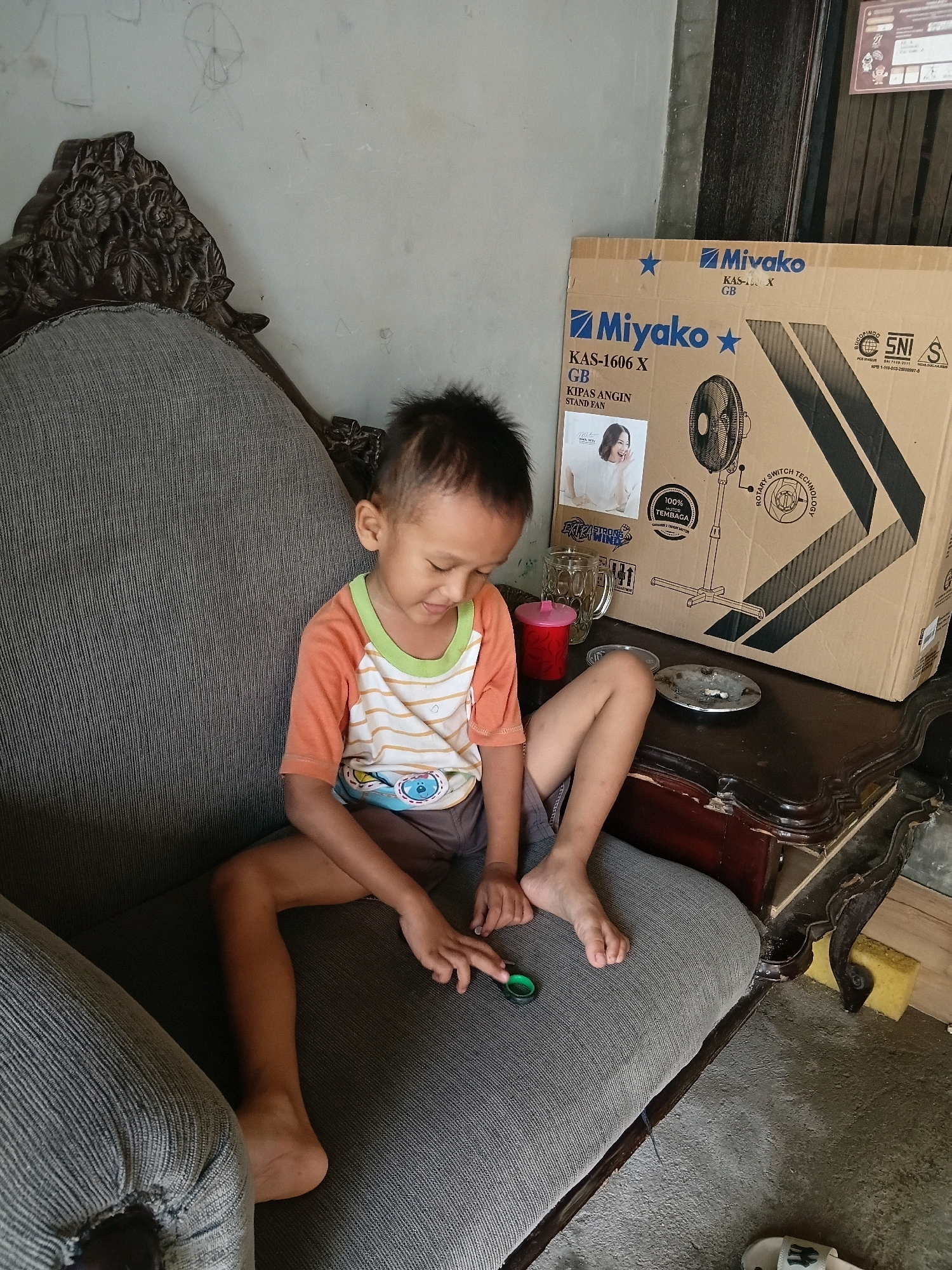 A young Indonesian boy with short black hair and light brown skin, wearing an orange and white striped t-shirt and grey shorts, is sitting on a grey upholstered sofa. He is looking down and playing with a small green object in his right hand. To the right of the sofa is a dark wooden table with a glass, a red cup, a metal ashtray, and a large cardboard box. The box has Japanese writing and an image of a fan on it. The room appears to be indoors, possibly in Cikal, Indonesia, with a concrete floor and a wall with some drawings. The lighting is natural and appears to be coming from the side.