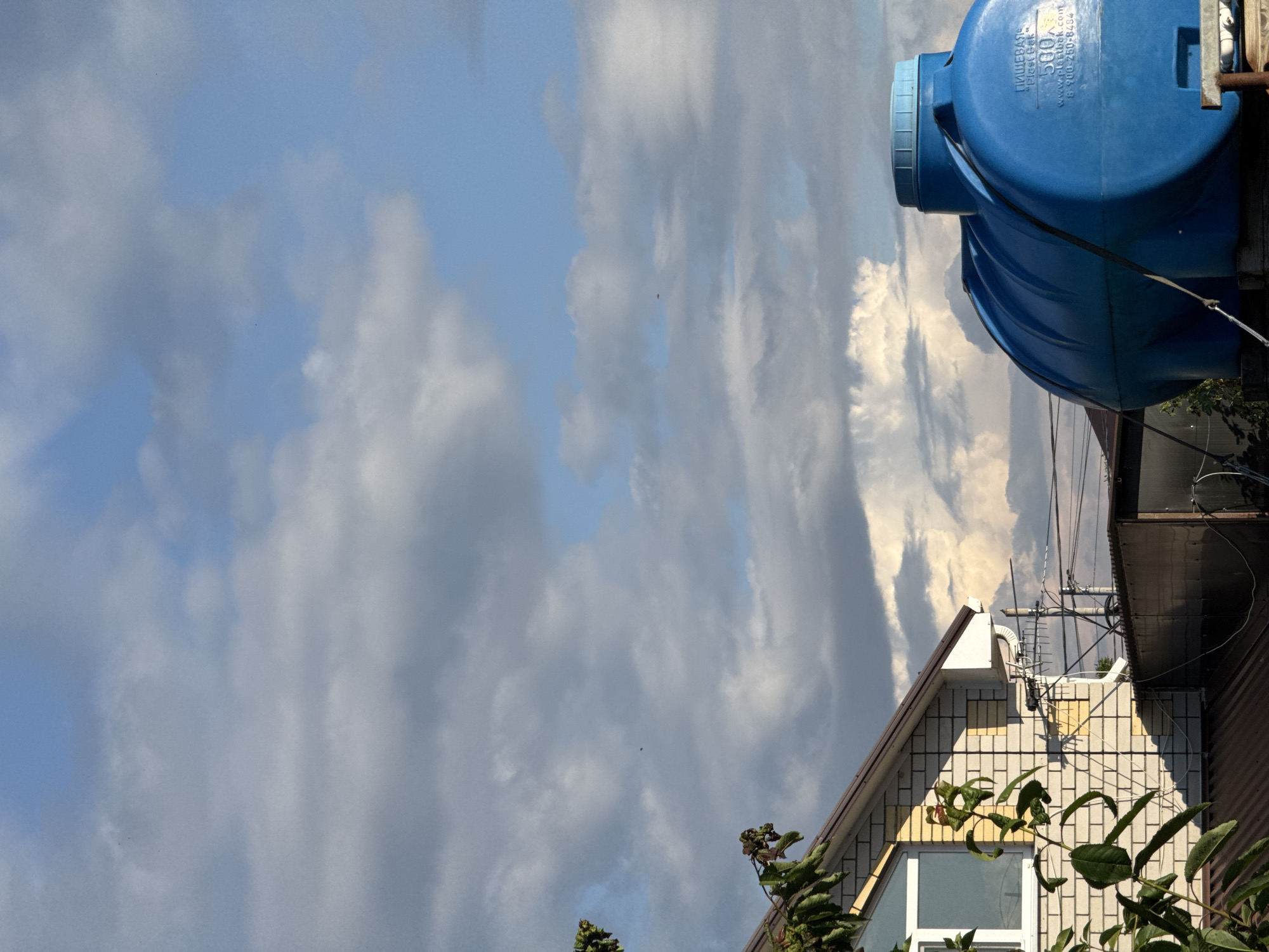 The image shows an outdoor scene with a bright blue plastic water tank prominently featured in the upper right. The tank has text visible on it, including "ПИЩЕВАЯ" (which translates to "food grade") and "PLASTEK" followed by "500 л" (indicating 500 liters). There are also web addresses and phone numbers. Below the tank, the roofline of a building is visible, constructed with light-colored brickwork and a dark-pitched roof. A window with a white frame is visible on the building. Lush green foliage from trees and bushes partially obscures the lower part of the building and extends into the foreground. The background is dominated by a dynamic sky with a mix of blue patches and dramatic, heavy clouds, suggesting varied weather conditions. The lighting indicates it is daytime, possibly late afternoon or early evening, given the shadows and the contrast between illuminated clouds and darker areas. The scene appears to be a residential or industrial setting in Mikhaylovsk, Russia. There are no people visible in the image. The overall impression is one of an everyday scene, perhaps documenting infrastructure like water storage against a backdrop of a somewhat moody sky.