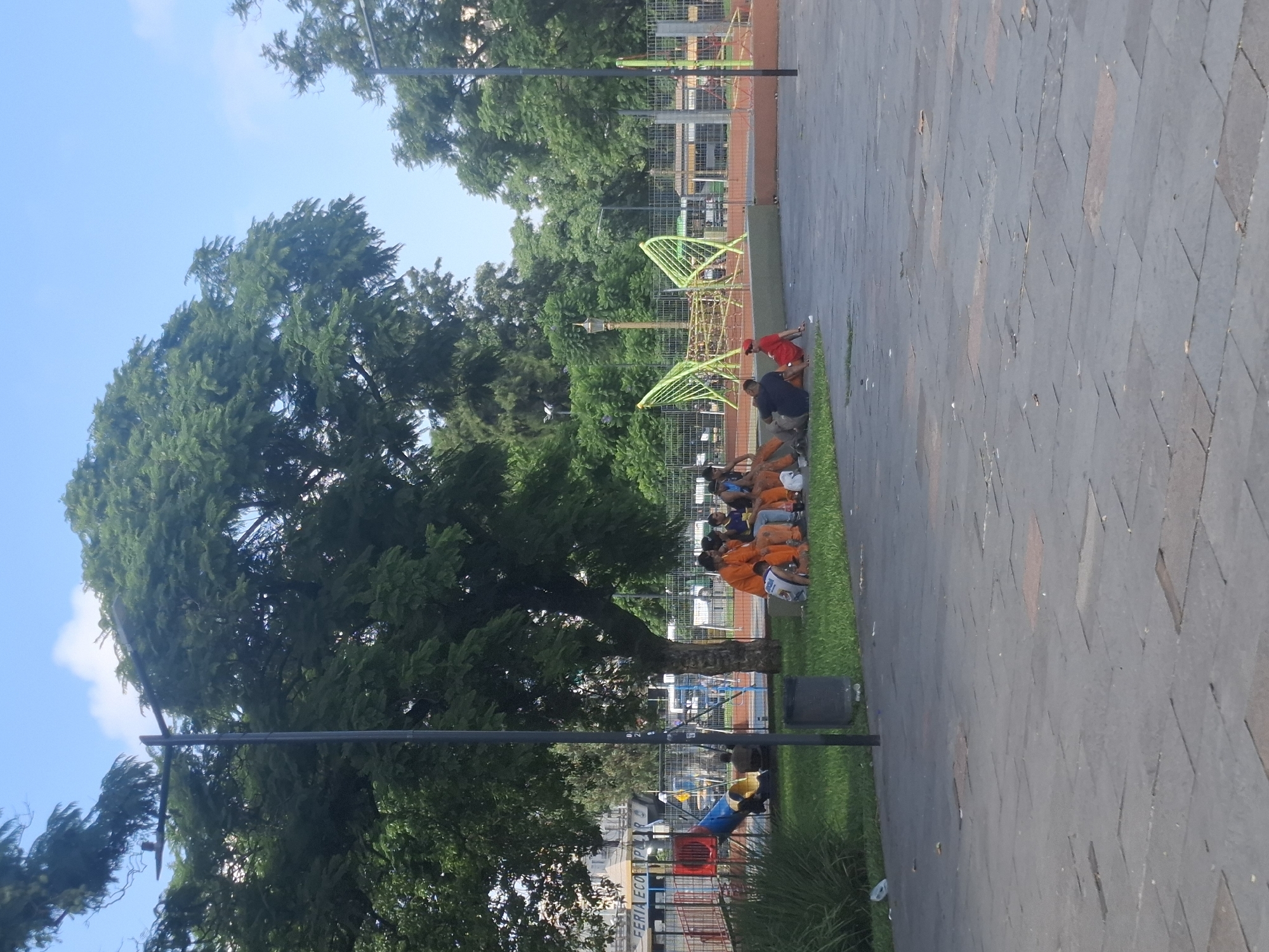 The image shows a scene in a park or public area, likely in Buenos Aires, Argentina, given the Latin American architectural style and coloring. A group of people, some wearing orange shirts, are sitting on the edge of a grassy area, adjacent to a paved walkway. There appears to be an adult male with the group. Beyond the people, a playground with green climbing equipment is visible. In the background, there are fences, trees, and possibly buildings. The sky is blue, suggesting a clear day. A lamp post is situated between the trees and grassy area.