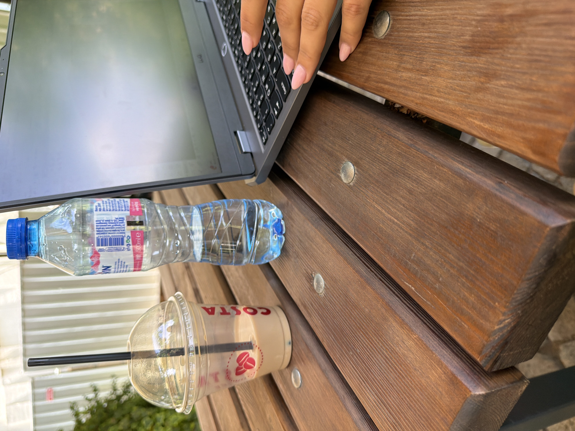 The image, taken from a high-angle perspective, captures a person's hands actively typing on a laptop keyboard outdoors. The individual has light pink manicured nails. The laptop, silver or grey in color, is open, with its screen reflecting a bright, possibly cloudy, sky.
Adjacent to the laptop on a sturdy, dark brown wooden surface, there are two beverages. A clear plastic bottle of "NATURA" brand water with a blue cap is visible, featuring a barcode and "500ml e" text on its label. Below it, a clear plastic cup from "COSTA" contains a light brown beverage, likely iced coffee, and a black straw. The "COSTA" logo, including two coffee beans, is clearly visible on the cup.
The items are placed on a wooden bench or table composed of planks with visible grain and metal fasteners. The background is softly blurred, revealing green foliage (bushes or trees) and a light-colored, possibly corrugated metal structure or fence. The bright natural lighting suggests a daytime setting. The scene depicts someone engaged in work or study in an outdoor environment, possibly a park or patio in Sofia, Bulgaria, combining productivity with refreshment.