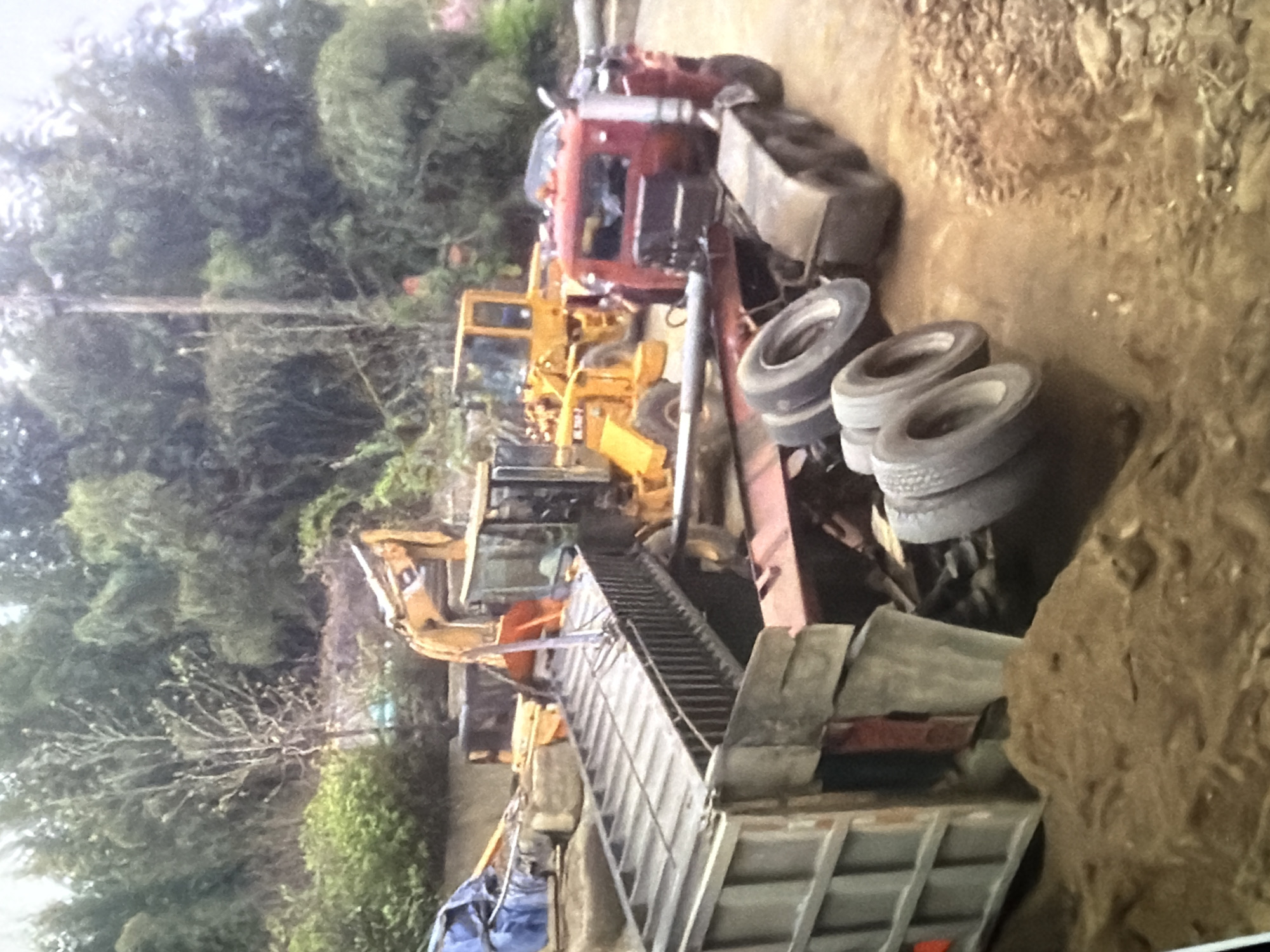 In an outdoor, dirt and debris-filled environment, a red tractor-trailer truck is overturned on its side, its trailer detached and its cab facing right. The trailer has a metal railing and appears to be a flatbed, with a long, empty metal structure on top. Several tires are scattered around the overturned truck, some still attached to axles. In the background, a yellow excavator is positioned behind the trailer, with its arm extended and holding a metal object above the trailer. Another yellow loader vehicle is partially visible to the left of the excavator. The scene is surrounded by trees and foliage.