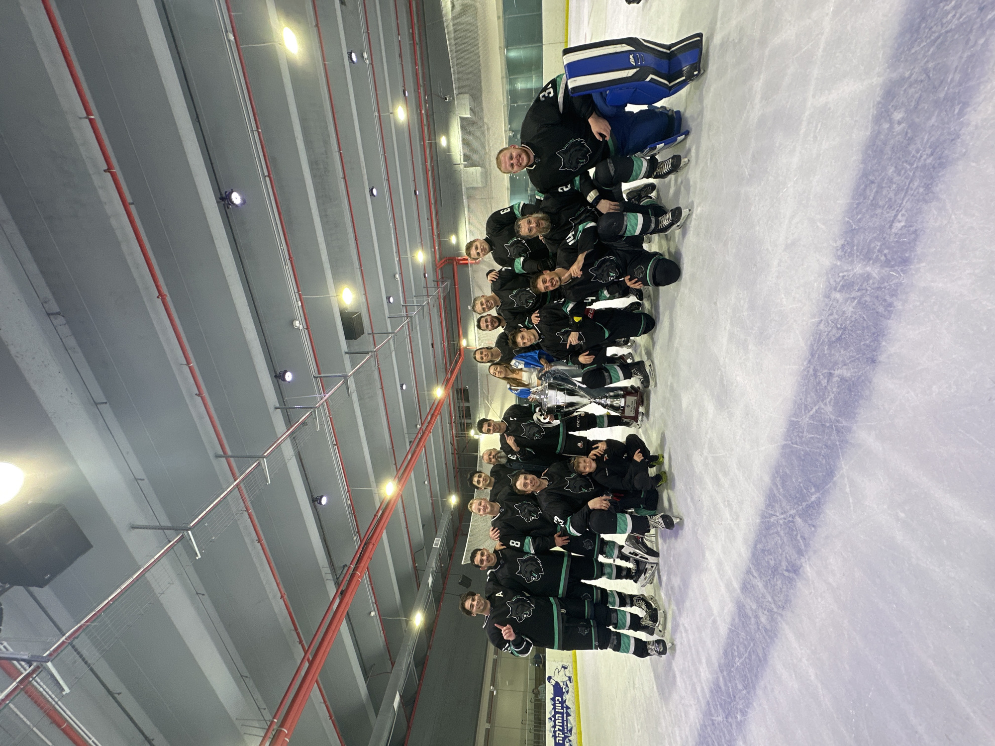 The image depicts a hockey team, predominantly males, posing for a photo on an ice rink. They are wearing matching black and green hockey uniforms with a wolf logo. Several individuals are holding a large trophy. The environment appears to be an indoor ice rink, with a concrete ceiling and industrial lighting. Visible in the background are signs, one of which reads "כיוון החלק" (Kivun Ha'Chalak), suggesting this is the Kivun Ha'Chalak Skating Arena located in Ashdod, Israel. The floor is an ice rink with visible skate marks. The lighting is bright, illuminating the scene.