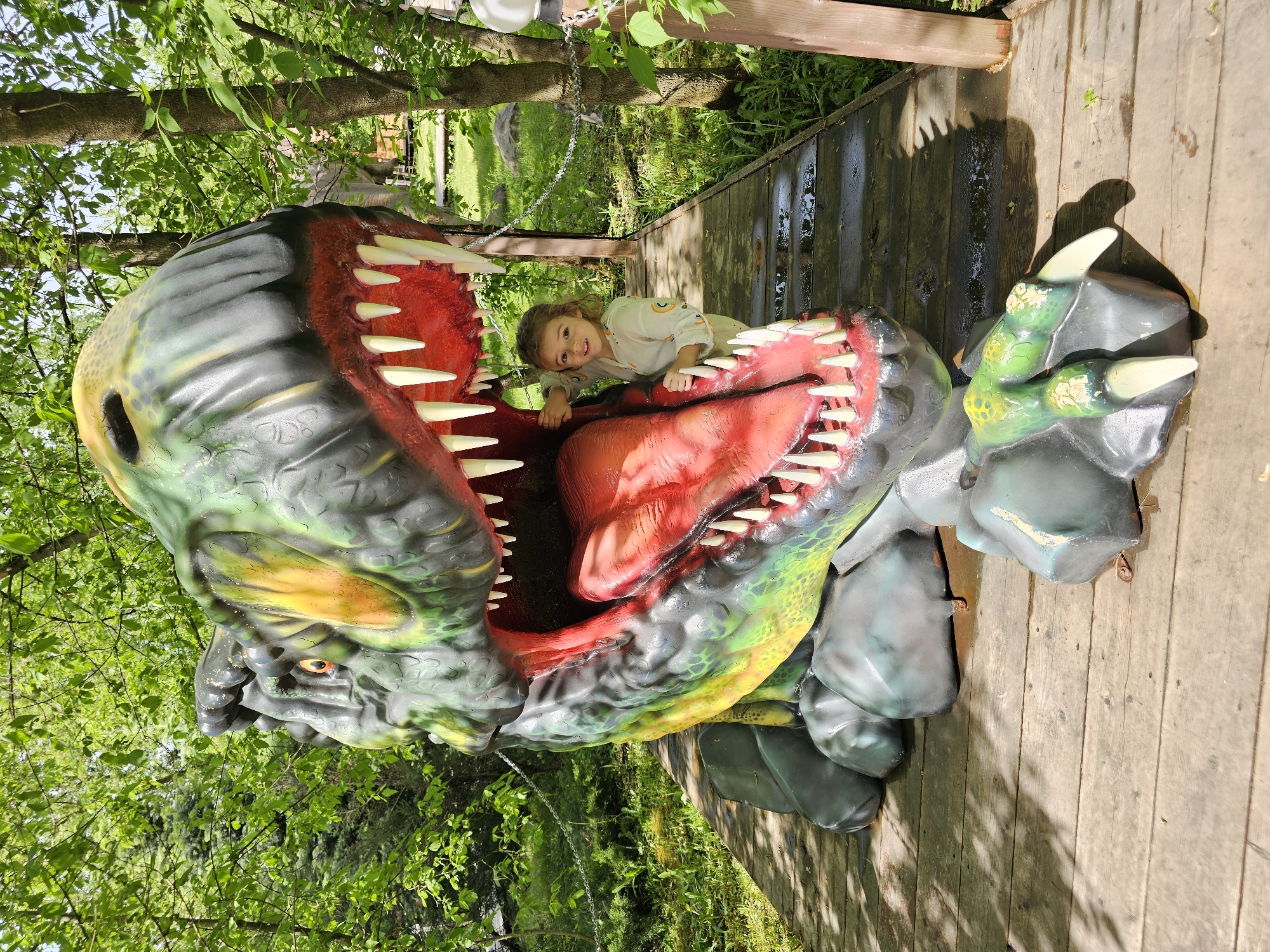 A girl is inside a large dinosaur head sculpture on a wooden platform. The dinosaur head is predominantly green, yellow, and black with a red interior mouth and prominent white teeth. The girl, positioned within the mouth, wears a light-colored shirt with rainbow-themed decorations. She is smiling and looking towards the camera. The scene is outdoors, surrounded by green foliage and trees, which provide partial shade. The wooden platform is wet in places. A chain is suspended in the background, likely a barrier or decorative element. The presence of the dinosaur head and the wooden platform suggest a recreational or park-like environment. The location in the city of Khimki, Russia, indicates a park or attraction within that city.