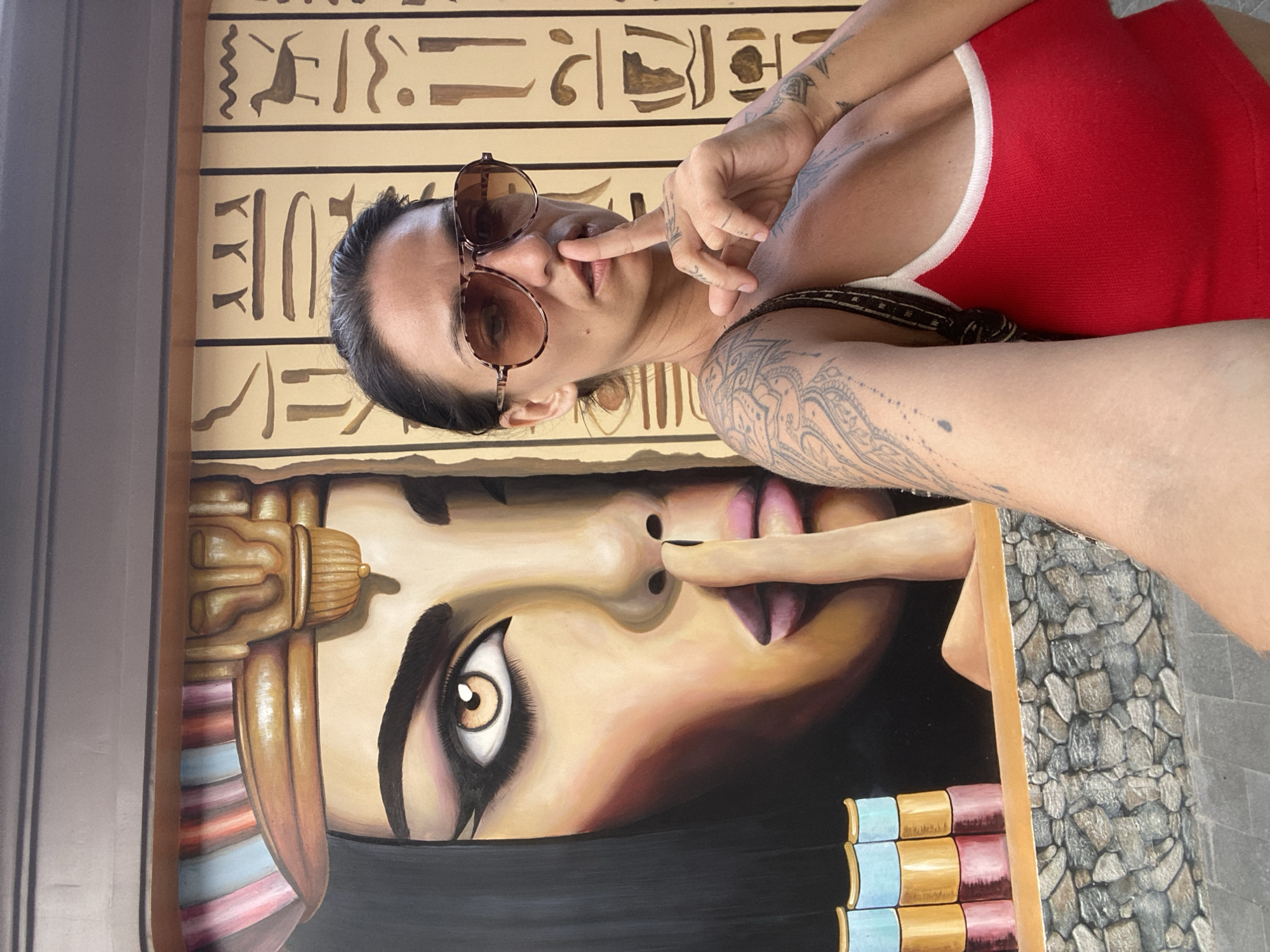 A woman with tattoos on her arms and chest stands in front of a mural. The mural depicts a stylized Egyptian queen with intricate headdress and hieroglyphics on the wall behind her. The woman is wearing large, tinted aviator sunglasses, a red tank top with white trim, and has her index finger pressed to her lips in a "shushing" gesture. The mural is painted on a rough, textured wall with a stone-like base. The image appears to be taken outdoors on a sunny day, with a hint of pavement visible in the foreground. The location is unknown, Vietnam.