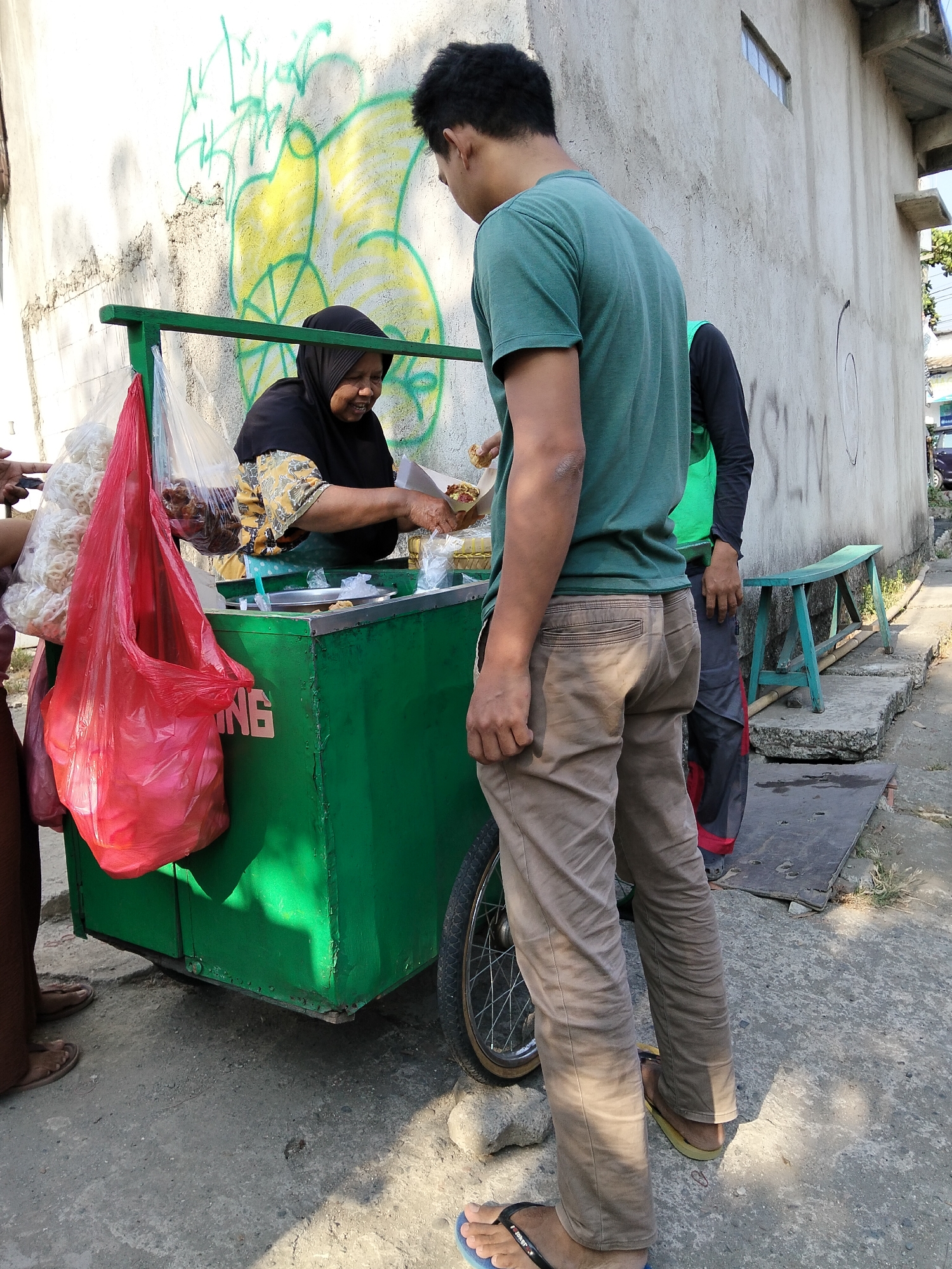 In the city of Kepandean, Indonesia, a street vendor is seen serving food from a green cart on a sunlit day. The cart is adorned with various food items and red plastic bags. A woman wearing a black hijab and a patterned blouse stands behind the cart, actively preparing food for a customer. Two men are standing in front of the cart, one is reaching out to receive food. The background features a textured wall with graffiti and a small window, hinting at an urban setting. The ground is paved with concrete, with some scattered rocks and a small green bench visible on the right side. The overall scene depicts a casual street food transaction in a local Indonesian community.