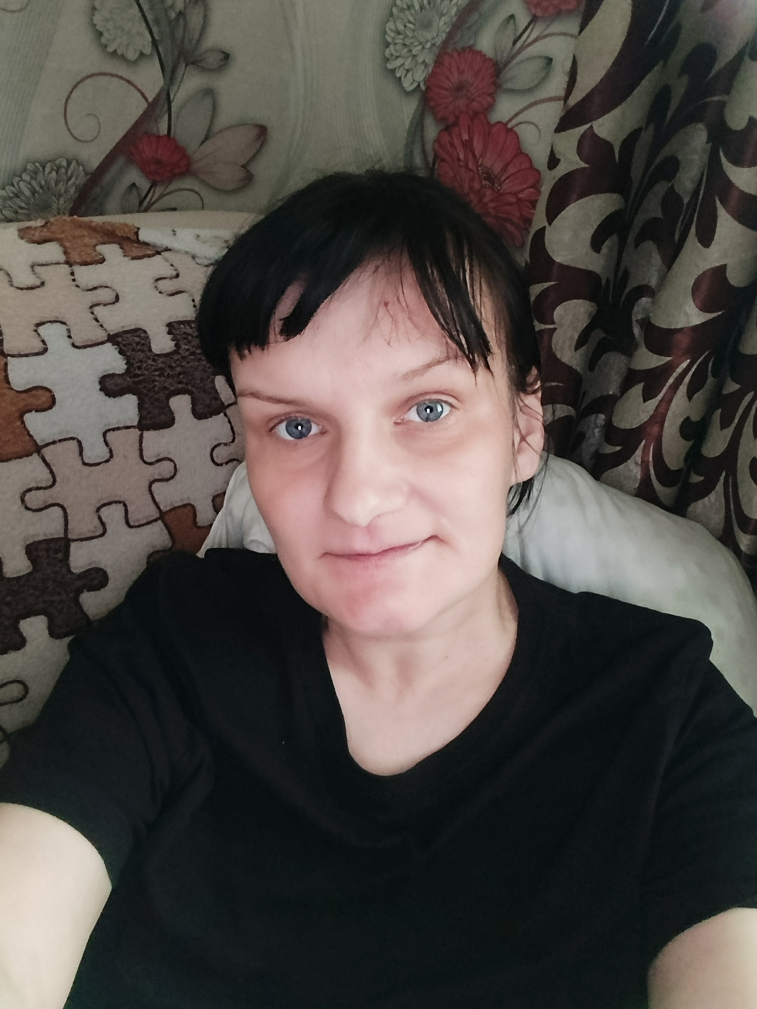 A woman with dark, short hair and blue eyes is pictured in a close-up, chest-up selfie, looking directly into the camera with a neutral to slightly smiling expression. She has bangs across her forehead, where a small reddish mark is visible. She is wearing a plain black t-shirt.
The woman appears to be indoors, reclining or leaning back against a surface. To her left, part of a fabric with a light and dark brown jigsaw puzzle pattern is visible, likely a cushion or blanket. Behind her and to her right, the background consists of patterned curtains or wallpaper featuring floral designs with red, grey, and white flowers and swirling foliage on a light background. A white pillow is partially visible behind her right shoulder and head. The lighting is soft and even, suggesting an indoor setting during the daytime. There is no visible text in the image.