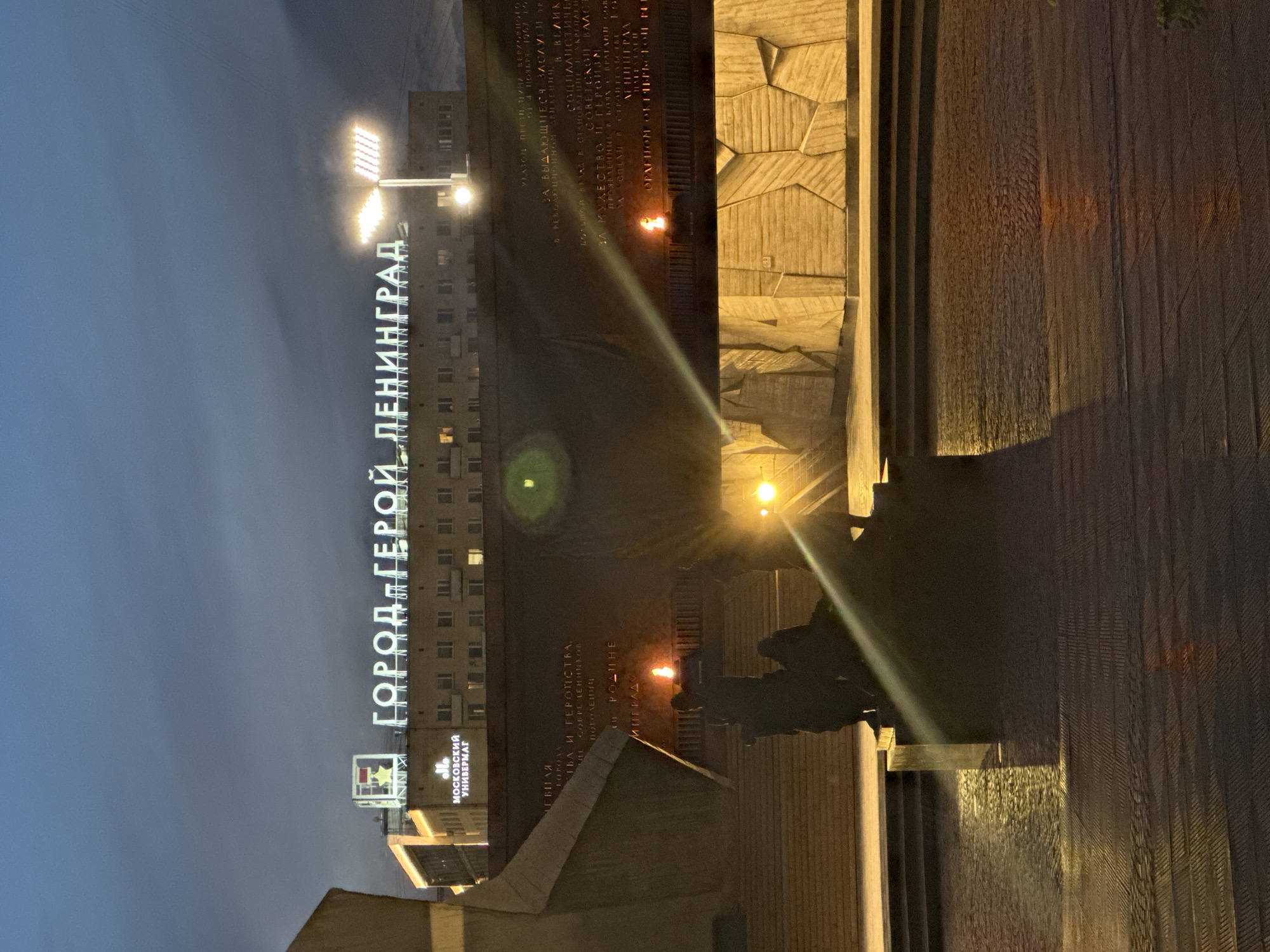 This nocturnal image captures the Monument to the Heroic Defenders of Leningrad at Pobedy Square in Saint Petersburg, Russia. The scene features monumental brutalist concrete structures under a dark, cloudy sky. In the lower-midground, multiple silhouetted sculptural figures, part of the monument, are visible on a tiered base. Two eternal flames burn with an orange glow, positioned centrally at the monument's base. The expansive, textured wall behind the flames displays dimly lit inscriptions in Cyrillic. The foreground ground plane consists of wet, reflective cobblestones and paving, mirroring artificial light sources. Prominent lens flares emanate from bright floodlights illuminating the scene. In the background, atop a multi-story building, large illuminated white letters spell "ГОРОД-ГЕРОЙ ЛЕНИНГРАД" (Hero City Leningrad). To the left of this main sign, another illuminated sign displays a red star emblem resembling a Soviet medal above the text "МОСКОВСКИЙ УНИВЕРМАГ" (Moskovsky Department Store).