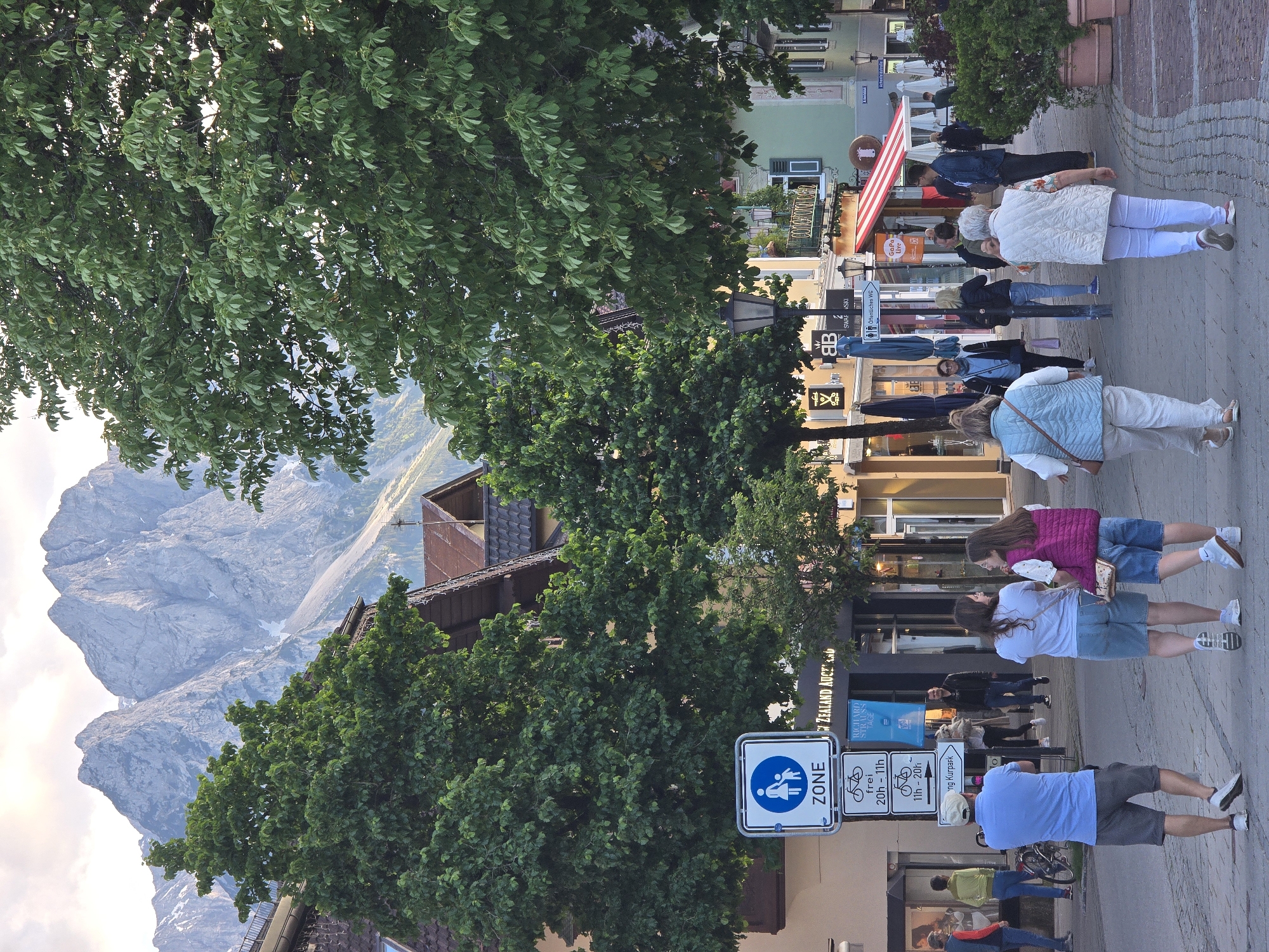 This detailed image captures a bustling pedestrian zone in Garmisch-Partenkirchen, Germany, with the majestic Bavarian Alps forming a dramatic backdrop under a partly cloudy, late afternoon sky. In the foreground, a paved street, constructed from light-colored pavers, is occupied by several individuals. On the left, a man in a light blue polo shirt, grey shorts, and a white baseball cap stands with his back to the viewer, holding a phone or camera. To his right, two young women walk side-by-side, engaged in conversation; one wears a white t-shirt and denim shorts, while the other sports a pink quilted vest over a white top and denim shorts. Further right and slightly ahead, two older women also walk away from the viewer; one wears a light blue quilted vest and light-colored pants, the other a white quilted vest and white pants. A black bicycle is parked on the left side of the street, near a lamppost. Prominently featured in the mid-ground are several street signs. A blue circular sign with a white pictogram of an adult and child indicates a "ZONE" (pedestrian zone). Below it, a white rectangular sign shows bicycle symbols with German text: "frei 20h - 11h" (free 8 PM - 11 AM) and "11h - 20h" (11 AM - 8 PM), likely indicating bicycle restrictions. Another sign points towards "Kurpark" (Spa Park) and identifies "Öffentliches WC" (Public Restroom). A banner for "RICHARD STRAUSS TAGE" (Richard Strauss Days) is visible, a cultural reference specific to Garmisch-Partenkirchen. The street is lined with multi-story buildings displaying traditional Alpine architecture, characterized by pitched brown-tiled roofs and numerous windows. Storefronts at street level include a "SWAROVSKI" shop, a partially visible "NEW ZEALAND AUCTIONS" sign, and "CORONADO CAFE" with a distinctive red and white striped awning. Several large, leafy green trees, likely horse chestnut, are interspersed along the street, their dense foliage partially obscuring buildings and framing the mountain views. Black ornamental lampposts provide street lighting, and large brown planters holding green shrubs are placed along the sidewalk. Various people populate the street in the mid-ground and background, walking, standing, or conversing. They are casually dressed, suggesting a warm and pleasant day. The overall atmosphere is relaxed and vibrant, indicative of a popular pedestrian area in a tourist town nestled within a stunning mountain landscape. The towering mountains in the distance are rugged and grey, with some visible snow patches and sparse vegetation, characteristic of