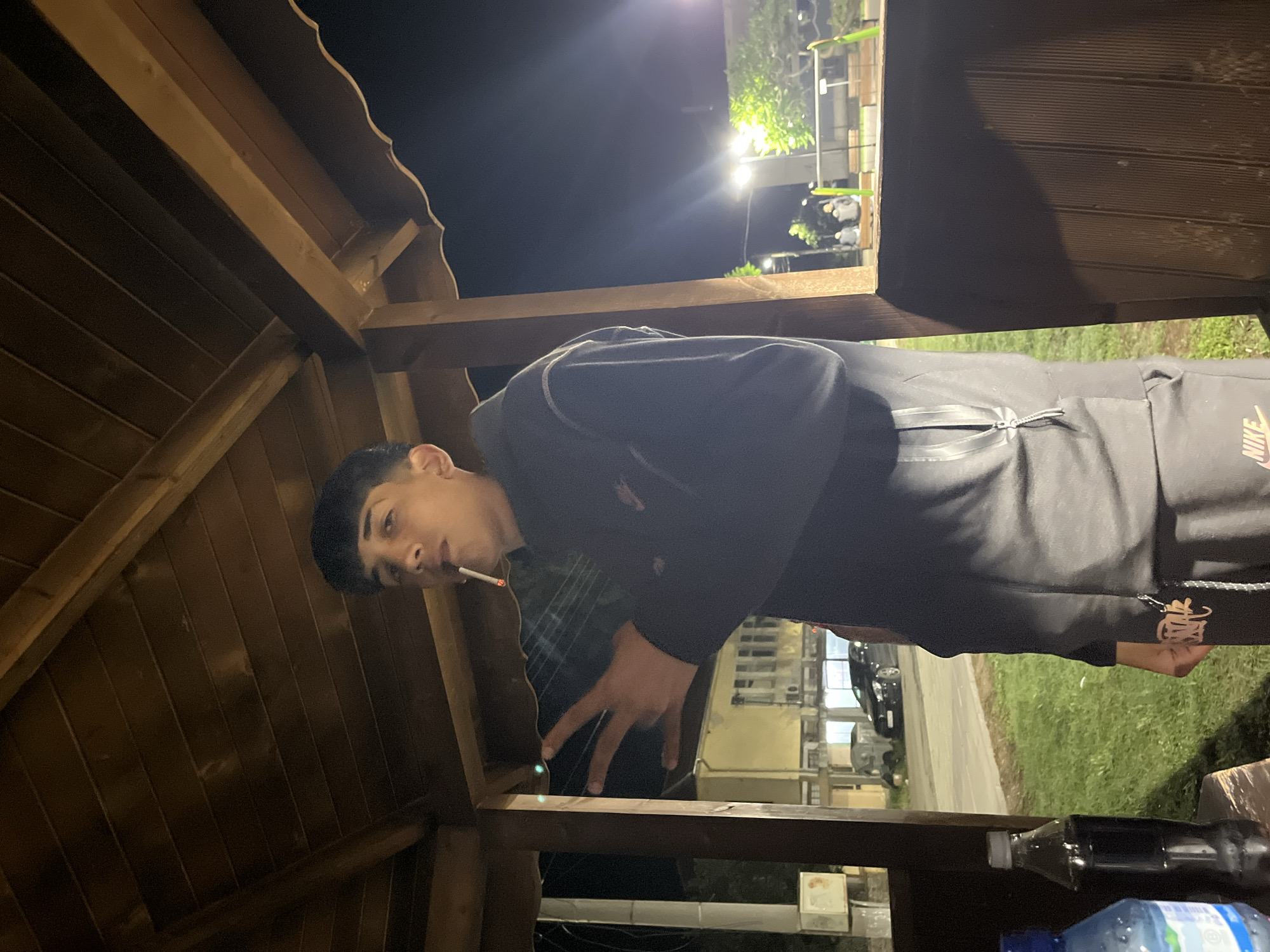 A young male is captured in a nighttime outdoor setting, positioned under a wooden gazebo or pergola structure. The image is oriented with the person's head to the top left and feet to the bottom right, suggesting the original shot was rotated.
The primary subject is a person, seemingly a young male, with dark hair, looking directly towards the viewer. He has a lit cigarette in his mouth. He is wearing a dark tracksuit, which features a prominent light-colored or gold-toned "NIKE" logo on what appears to be the right pant leg or sleeve. His right hand (from the viewer's perspective) is raised, with two fingers extended in a V-sign or peace gesture.
The setting appears to be a casual outdoor area at night. The wooden structure has a slatted roof and visible support beams. Beyond the shelter, the background is dark, illuminated by several bright artificial light sources that create lens flares. These lights highlight some green foliage and indistinct distant structures or buildings. A paved road and a patch of grass are visible on the right side of the frame, with a dark car parked further down the road. In the foreground, on a surface within the wooden structure, two plastic bottles are visible: one dark-colored, possibly a soda, and one clear/blue, likely a water bottle. The overall lighting is dim in the foreground, contrasting with the bright background lights.
The location context is Lozarevo, Bulgaria.