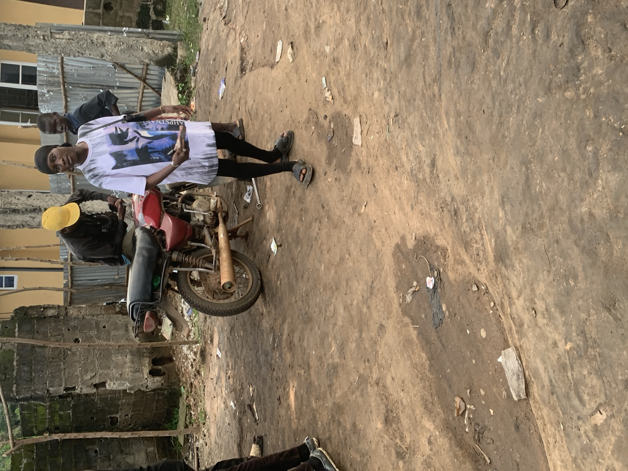 A woman, wearing a white t-shirt with a graphic print and black leggings, stands in the foreground of a dirt yard in Shagamu, Nigeria. She is wearing grey slip-on sandals and has a black headscarf. To her left, a man wearing a yellow cap and dark clothing is partially visible, leaning over a red motorcycle. Behind the woman, a young boy in a black shirt stands looking towards the camera. The environment appears to be a disused or semi-abandoned area with a damaged concrete structure on the left and corrugated metal fencing in the background. The ground is uneven and covered in dirt, with scattered debris including what appears to be plastic bags and other refuse. The overall scene depicts a casual outdoor gathering in a somewhat dilapidated urban setting.