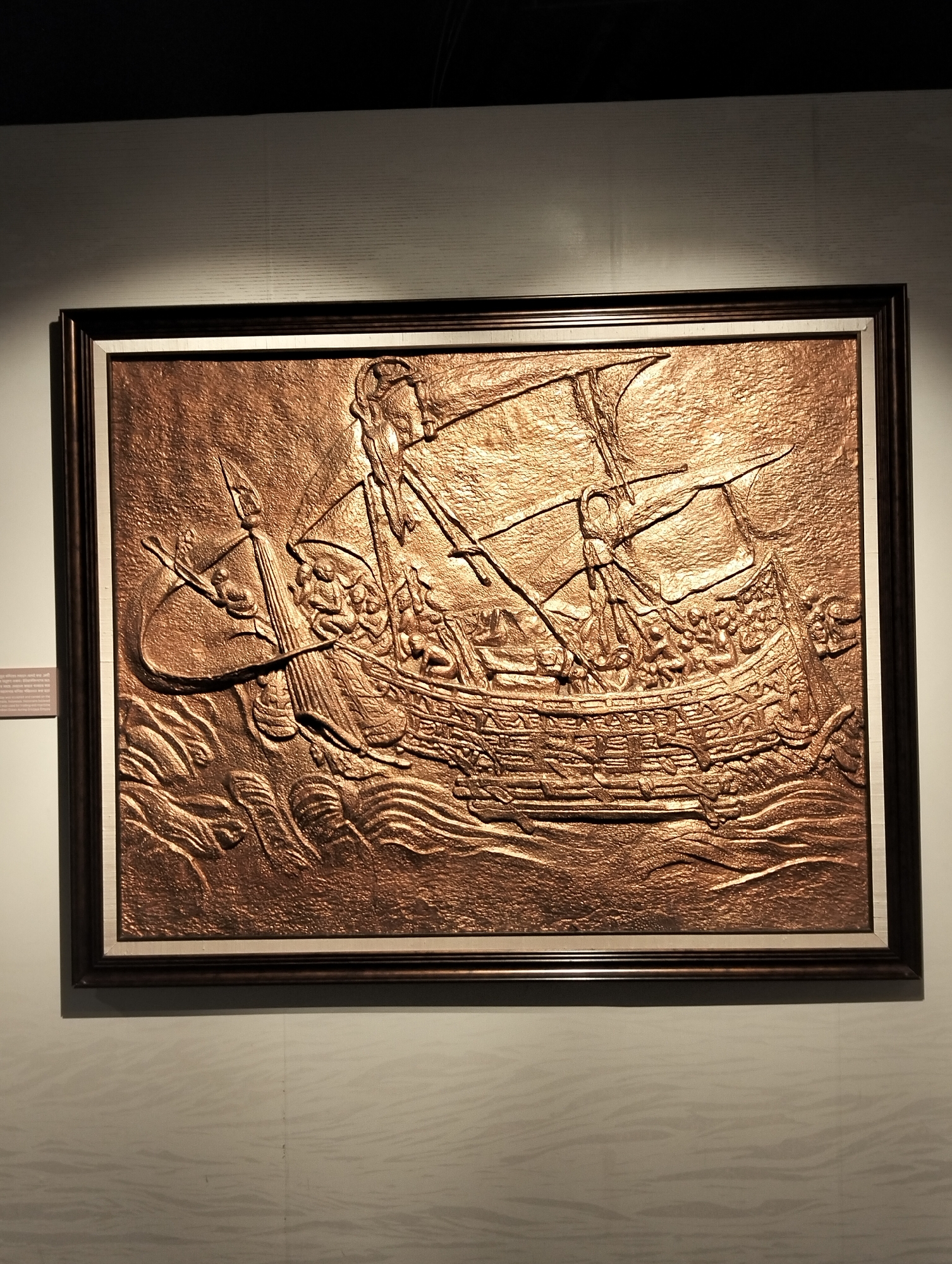 The image features a prominent, rectangular, framed relief sculpture crafted from a metallic material, likely copper or bronze, given its rich, reddish-brown hue and reflective surface. The sculpture depicts a historical sailing ship, possibly a galleon or a similar large vessel, navigating through turbulent waters.
The ship is shown in profile with multiple sails, some appearing full, suggesting movement. Numerous human figures are intricately detailed on the ship's deck. These stylized figures appear to be crew members or warriors, some seemingly engaged in tasks or holding objects that might be weapons or tools. One figure is prominently positioned at the bow, looking forward, while another seems to be steering at the stern. The sea beneath the ship is rendered with strong, undulating waves, emphasizing the dynamic nature of the scene.
The artwork possesses a textured, hammered surface that catches the light, creating dramatic highlights and shadows that enhance its three-dimensional depth. It is enclosed within a dark, possibly wooden, frame with a contrasting lighter inner border. The sculpture is well-lit, drawing attention to its intricate details.
The artwork is mounted on a light-colored wall. To the left of the frame, a small rectangular plaque is visible. Although the text is largely illegible, faint words suggest it might provide information about the artwork's subject, potentially linking it to "Dhaka," indicating its historical or cultural relevance to the region of Dhaka, Bangladesh. The top part of the image shows a dark ceiling or overhead structure.