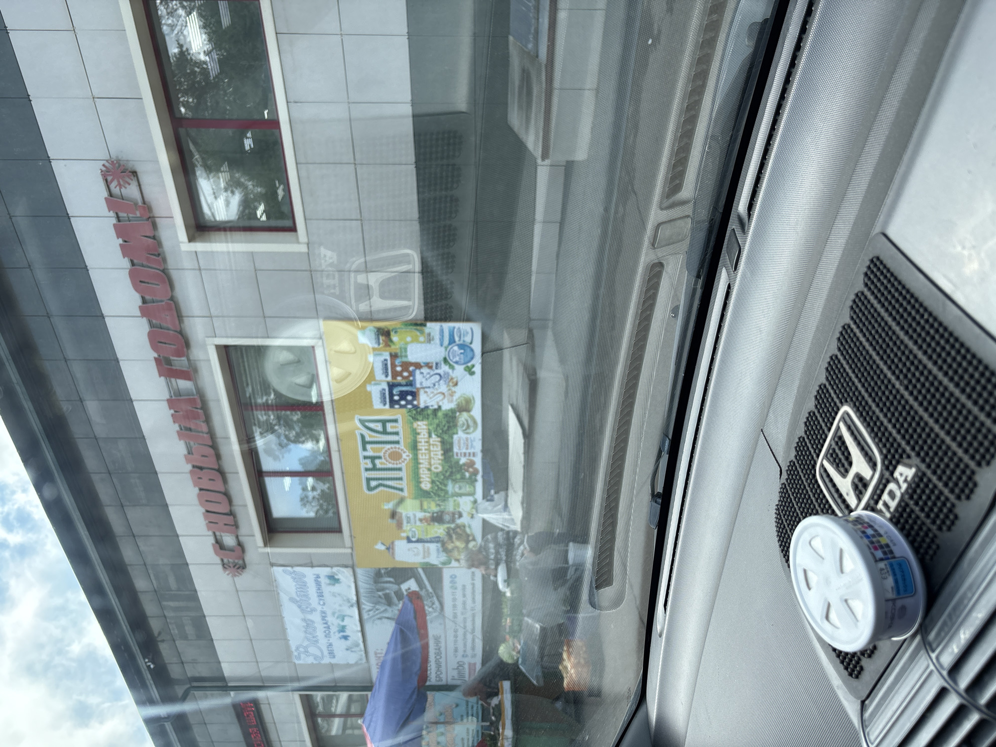 This image captures a street-level view through a car windshield. The foreground shows the dashboard of a car, featuring a white circular air freshener and a black mat with a silver Honda logo. The mid-ground and background reveal the exterior of a building with a tiled facade. A large red sign above the building reads "С НОВЫМ ГОДОМ!" (Happy New Year!). Below this, a banner displays the word "ЯНТА" in large yellow letters, followed by smaller text and images of various products. To the left of the banner, a market stall is visible under a blue and red umbrella, with people attending to produce. The overall environment suggests a commercial area within a city. The sign with "С НОВЫМ ГОДОМ!" is a common decoration during the New Year's holiday season in Russia, and the presence of a market stall indicates a public space. Based on the available information, this scene is likely located in the city of Bratsk, Russia, where similar commercial setups and holiday decorations are prevalent.