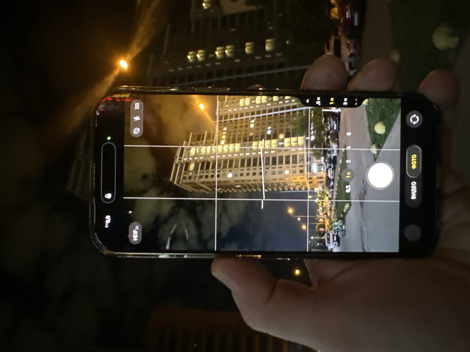 A person is holding an iPhone with its camera app open, pointed at a building at night. The iPhone camera screen shows a tall building with many illuminated windows. A street with parked cars is visible to the right. The buildings appear to be located in Kazan, Russia based on the architectural style of buildings visible in the background. The environment is dark.