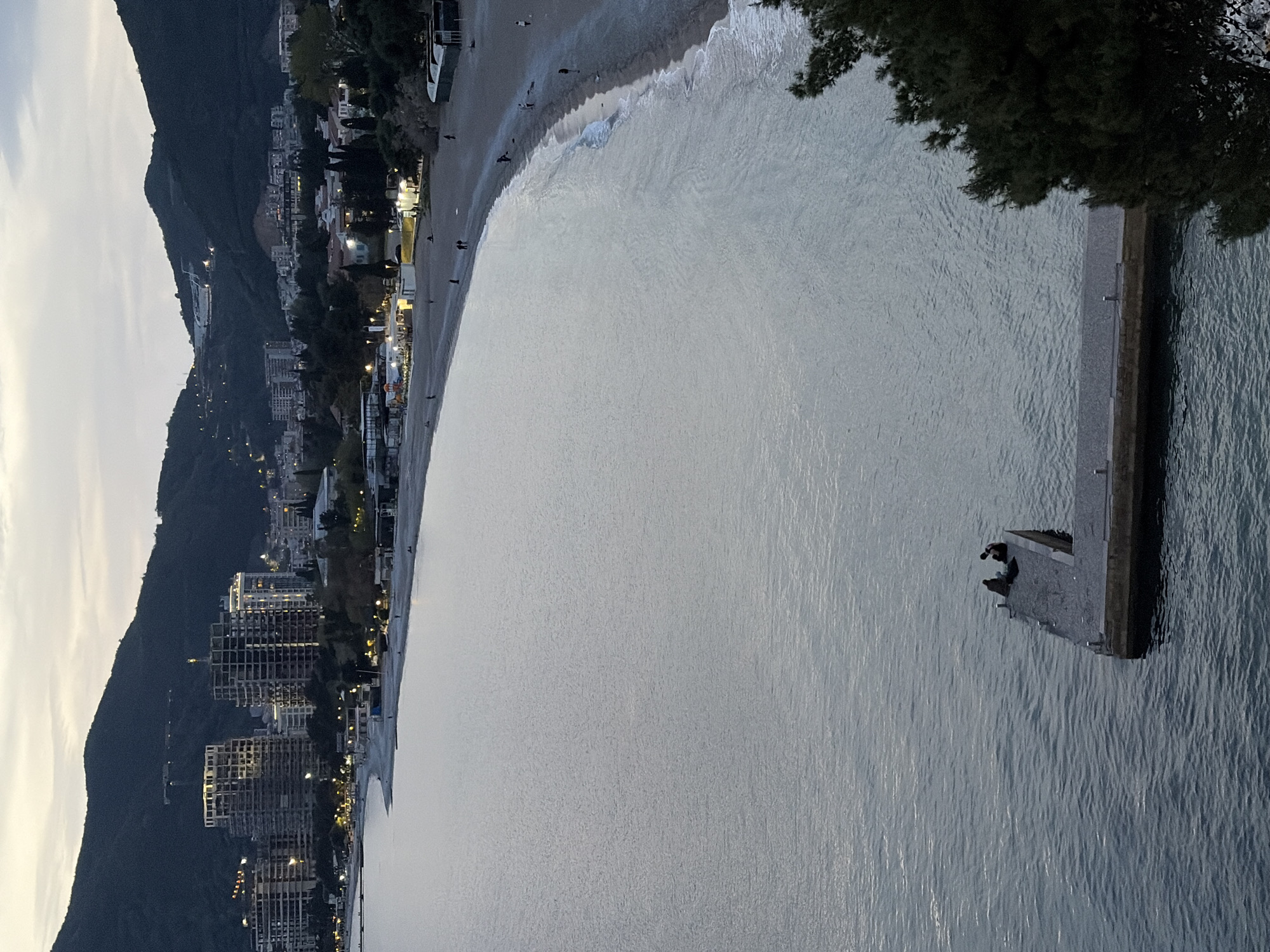 The image shows a coastline in Budva, Montenegro, at dusk. The Adriatic Sea occupies most of the frame, with a concrete pier extending into the water on the right side. Two individuals are seated on the end of the pier. A beach curves along the coast, lined with buildings and vegetation. Mountains rise in the background on the left, with city lights visible.