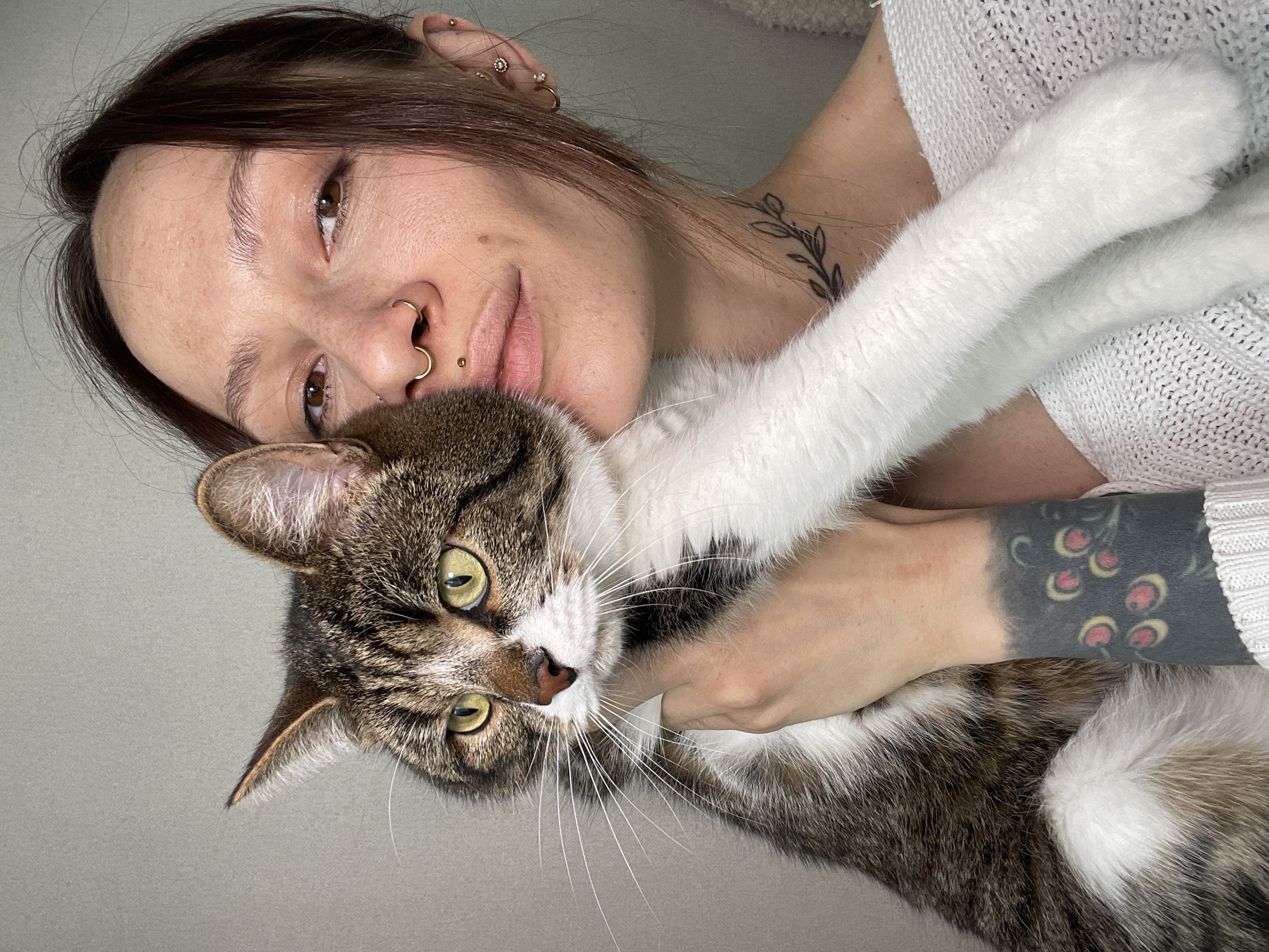 A fair-skinned individual with dark hair is holding a tabby cat. The individual has multiple piercings in their ear and nose and visible tattoos on their neck and arm. The cat has green eyes and white markings on its face and paws. The background is a light neutral color. There is no information to indicate the location of this photo as Pervomaysky, Russia.