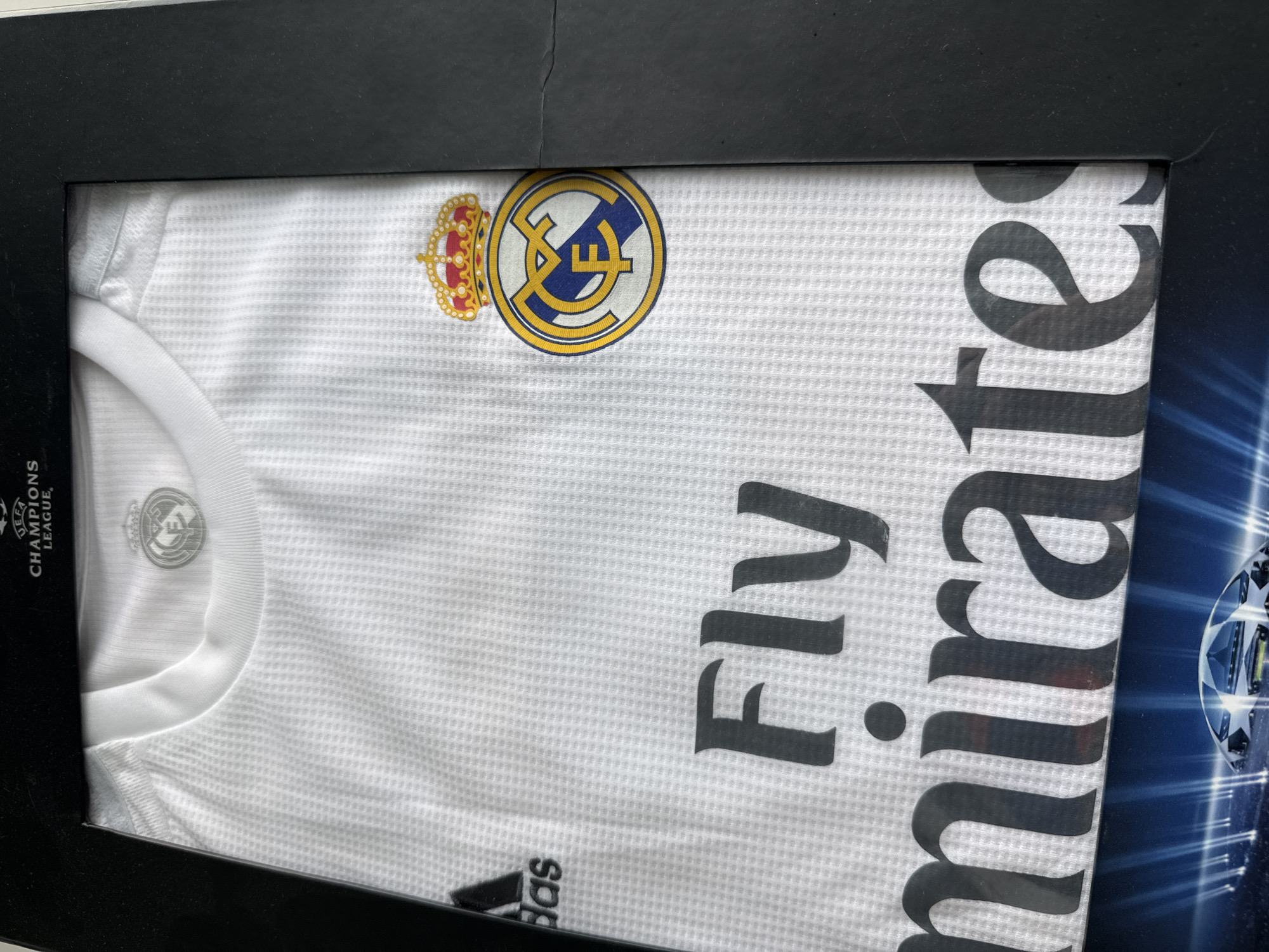 The image shows a framed Real Madrid soccer jersey. The jersey is white with the Real Madrid crest, the Adidas logo, and the sponsor "Fly Emirates" printed on it. The frame appears to be black. The image does not contain sufficient information to confirm a location in Vladivostok, Russia. No people, actions, or interactions are visible.