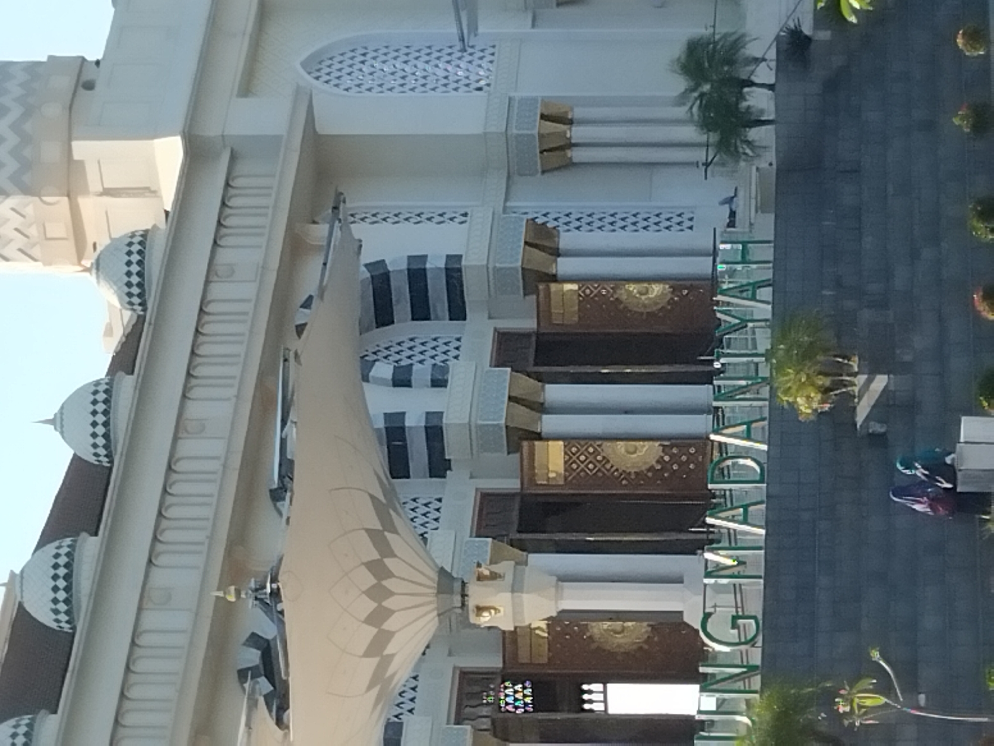 The image captures a mosque with a sign that reads "UNG MADANIYAH" in green, illuminated lettering. The mosque features white walls with decorative arches and patterned windows. Several domes are visible on the roof. In front of the mosque, there is a tiered stone structure with some greenery and palm trees. Two women in hijabs are seated on the stone steps in the foreground. The scene appears to be located outdoors, with a clear sky above.