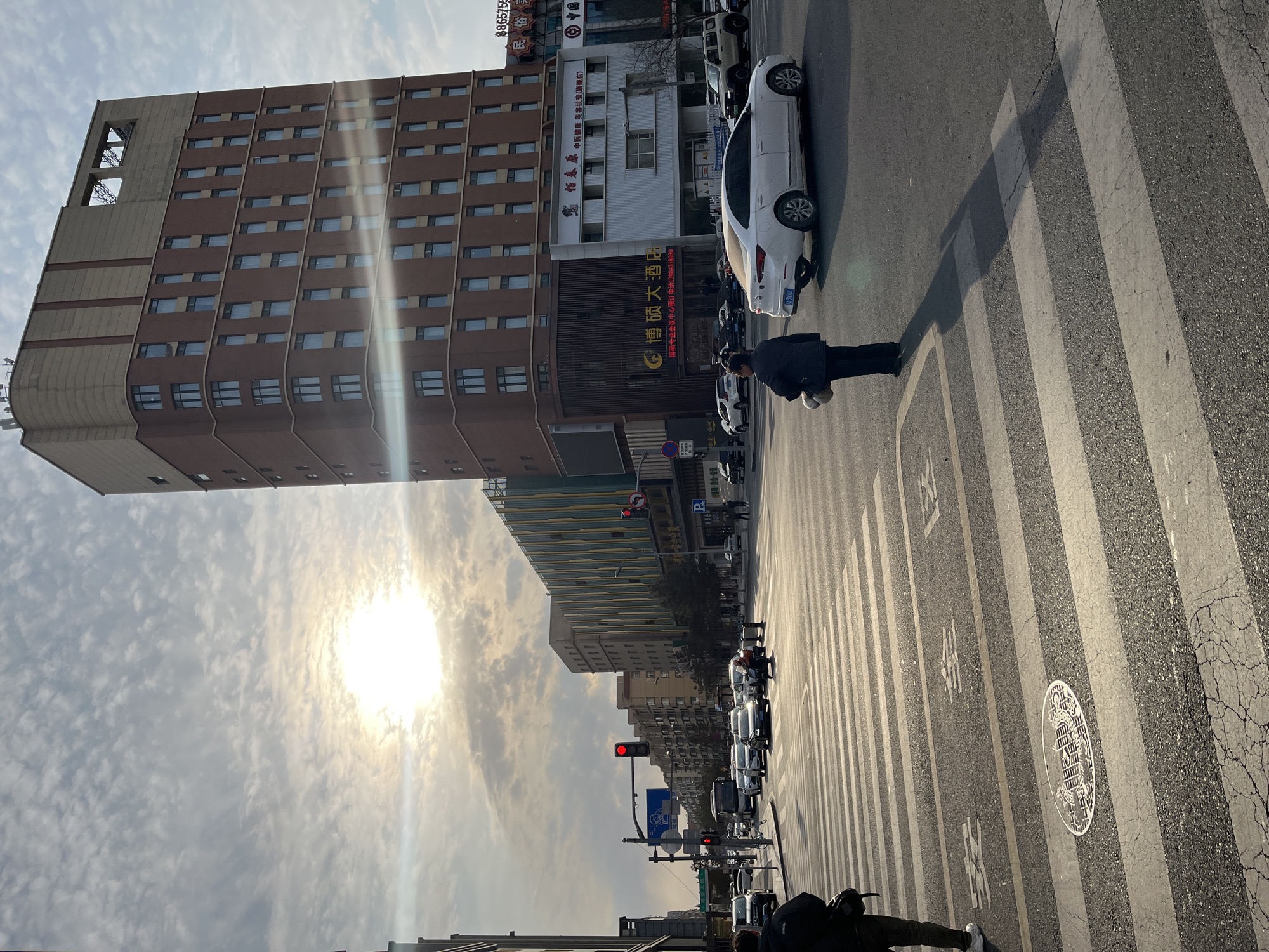 The image shows a street scene in Changchun City, China. A tall, brown building occupies the left side of the frame, partially obscured by clouds and the sun. In front of this building, there are other buildings. Cars and other vehicles are visible, including a white car parked on the right side of the street. A person is walking across a crosswalk in front of the building. A traffic light with a red light is visible in the distance. A sign that says “博硕大酒店” is visible on one of the buildings.