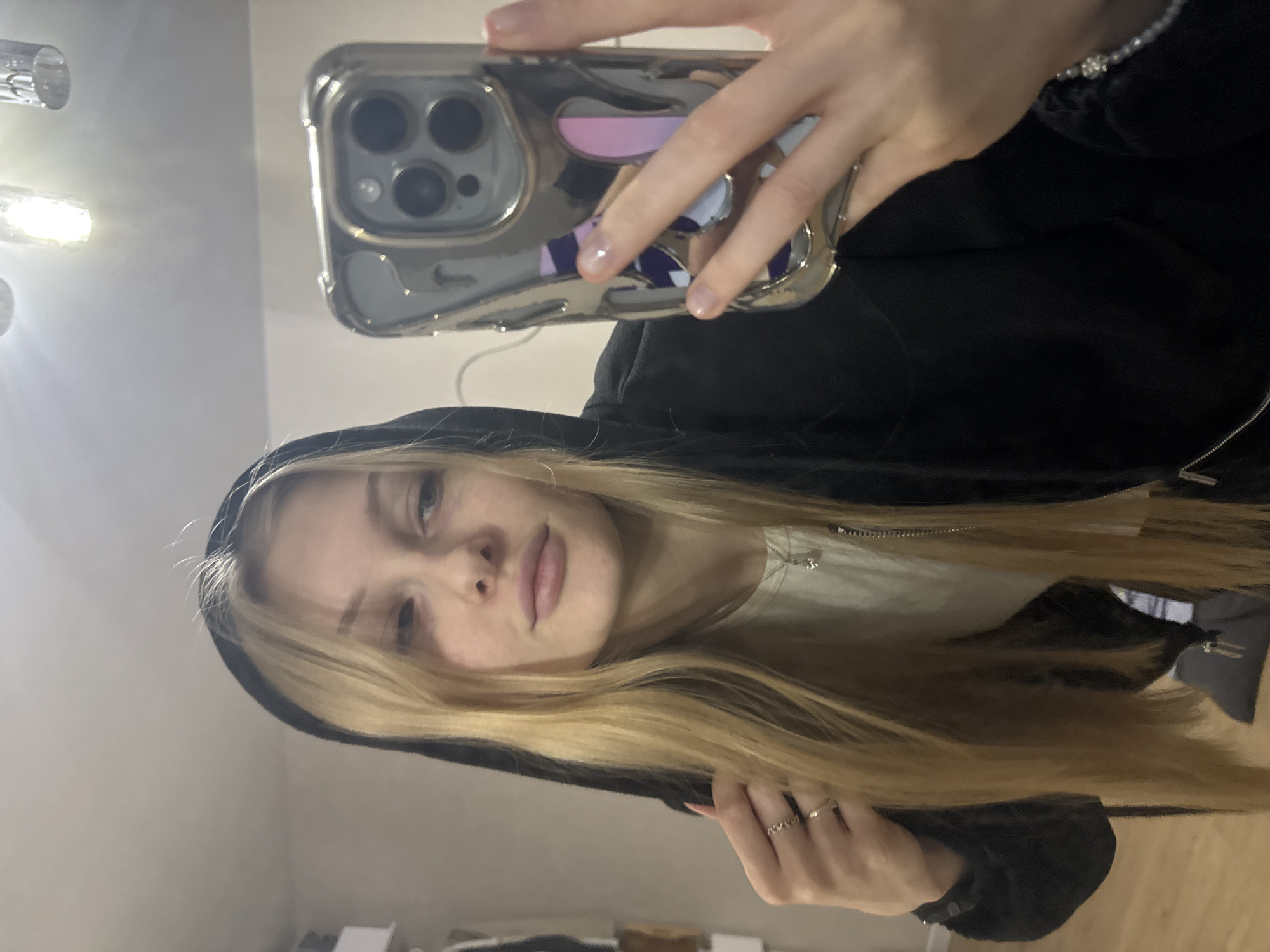 The image features a young woman taking a selfie indoors. She has long, straight blonde hair, light-colored eyes, and a neutral, calm expression. She is wearing a black hooded garment, possibly a hoodie or jacket, over a light-colored (likely white) t-shirt. On her right hand, several rings adorn her fingers, and a beaded bracelet is visible on her left wrist.
The woman holds a smartphone in her left hand, positioned prominently in the upper right portion of the frame. The phone is encased in a shiny, metallic, chrome-like case that features a distinctive cut-out design on the back. The phone's triple camera module is clearly visible through the case. Reflections within the glossy phone case reveal elements of the background, including patches of purple and white patterns.
In the upper left corner of the image, a bright, multi-bulb ceiling light fixture is visible, casting a strong glare. The rest of the background is an indistinct indoor setting, with light-colored walls and hints of furniture, suggesting a casual home environment. The image is presented in a tilted orientation, with the woman's head positioned towards the left side of the frame. The specific location is Perm, Russia. No visible text is present in the image.