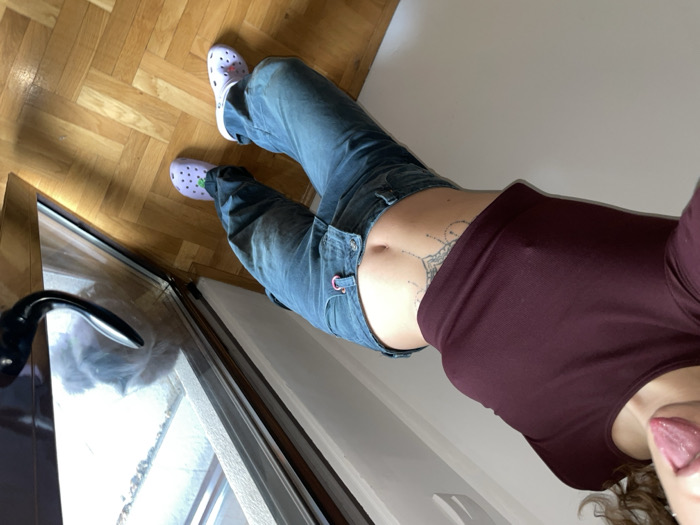 The image presents an interior scene located in Boreti, Montenegro. A female individual is shown in a low-angle selfie, capturing her torso and lower body. She is attired in a form-fitting, long-sleeved maroon crop top, which reveals her midriff and navel, displaying a belly button piercing and an ornate, mandala-style tattoo on her lower abdomen. Nipple prominence is visible through the fabric of the top. She wears loose, light blue denim-style pants with a visible metal ring on the left side of the waistband, and light purple Crocs-style clogs adorned with white dots and green strap accents. Her mouth is open with her tongue extended. The immediate environment includes a polished wooden parquet floor, a plain white wall, and a glass door or large window on the right. Beyond the glass, a blurred exterior view shows a light-colored surface and a grey, furry object, consistent with a curled-up cat, positioned near the bottom of the glass.