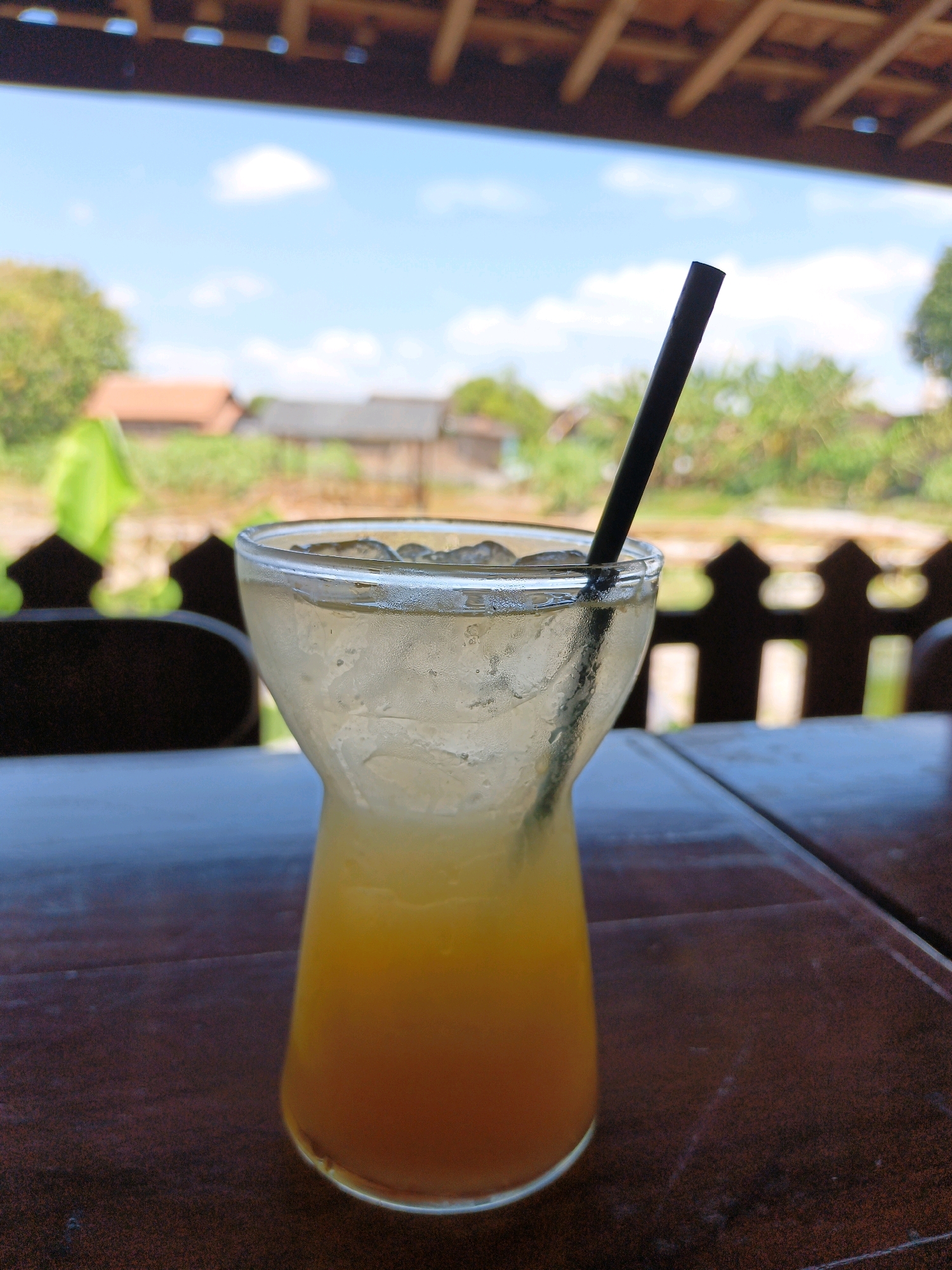 A close-up shot showcases a tall glass filled with a layered beverage, featuring a vibrant orange hue at the bottom fading to a lighter yellow at the top, topped with ice and a black straw. The glass rests on a dark brown wooden table, the surface of which is slightly worn. In the background, a blurred outdoor scene reveals a wooden railing with decorative cutouts, beyond which lies a landscape of greenery and rustic buildings under a bright blue sky with scattered clouds. The overall setting suggests an outdoor seating area, possibly a cafe or restaurant, in Jonggrangan, Indonesia, where the ambient light indicates daytime.