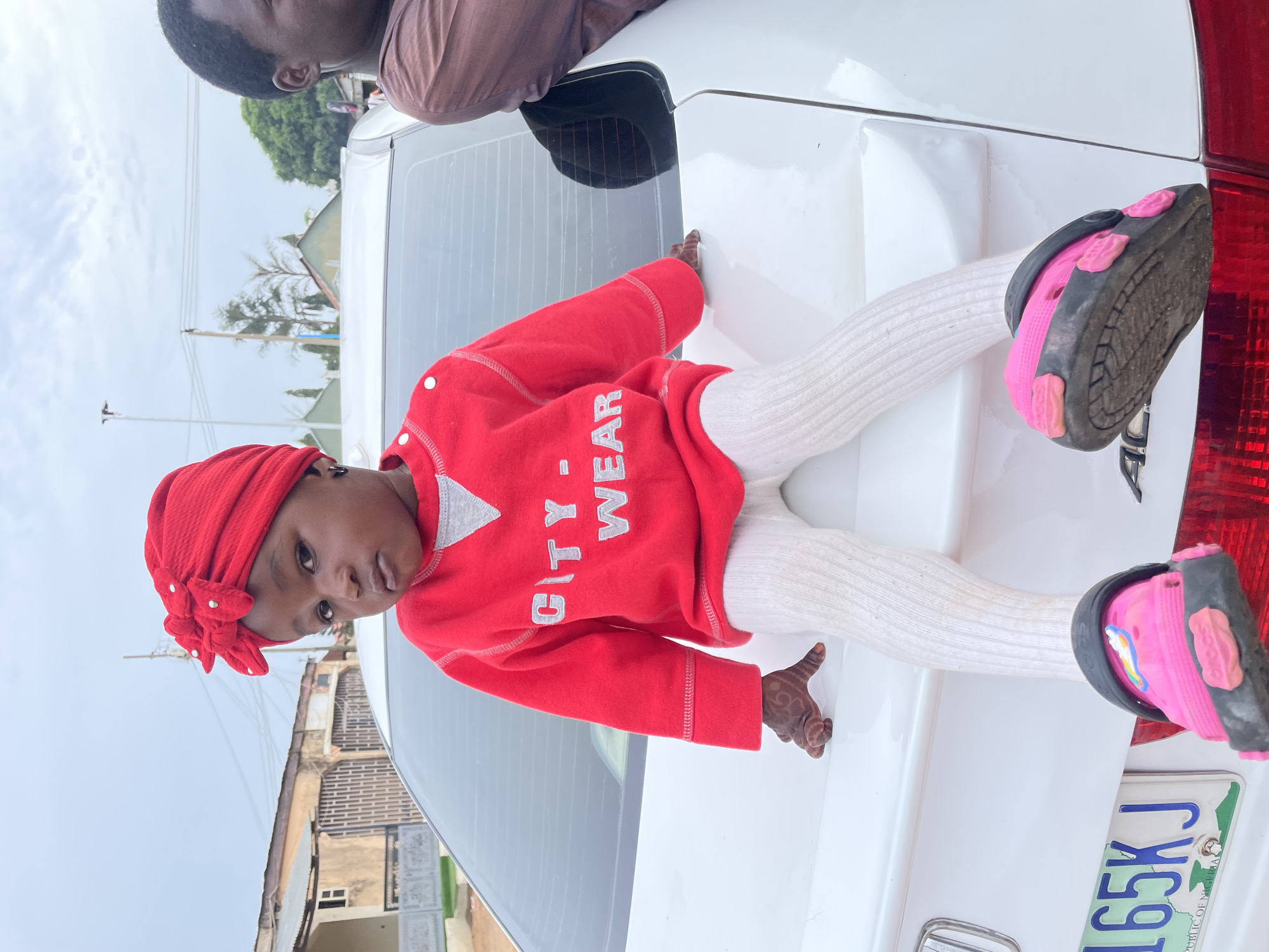 A young child in a red turban, red sweater with "CITY WEAR" printed on it, and white leggings, is sitting on the open trunk of a white car. The child is wearing pink and black Crocs. The car's license plate is partially visible and reads "165KJ," with the word "NIGERIA" below. A man in a brown shirt is visible in the upper portion of the image. The location appears to be a residential area in Barakin Akawo, Nigeria, with buildings visible in the background. The sky is overcast.
