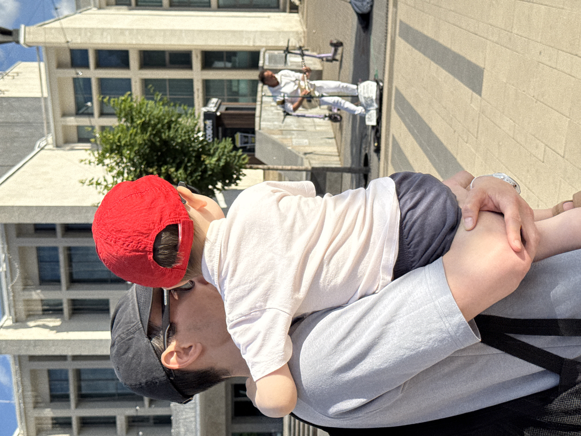 The image depicts an outdoor urban scene in the city of Izhevsk, Russia, on a bright, sunny day.
In the immediate foreground, an adult male holds a young child. The adult, viewed from behind and the right side, wears a black baseball cap, dark sunglasses, a light grey short-sleeved t-shirt, and has a black backpack strap visible over his left shoulder. A silver watch or bracelet is present on his left wrist. The child, held with its back to the camera and facing towards the right, wears a red baseball cap, a light pink or off-white short-sleeved polo shirt, and dark blue shorts. The child's left arm is wrapped around the adult's neck/shoulder.
In the midground, slightly to the right, a male street musician is performing. He is dressed entirely in white, wearing a hooded sweatshirt and pants. He stands on a white self-balancing scooter (hoverboard) and plays a light-colored acoustic-electric guitar. A small white collection box or sign is on the ground in front of him. Behind the musician, two purple electric scooters are parked on the paved surface.
The background features large, multi-story buildings with light beige facades and numerous rectangular windows arranged in a grid pattern with dark frames, characteristic of modern or post-Soviet architecture. A small green leafy tree stands between the foreground figures and the buildings. Above the tree, a dark rectangular sign with white Cyrillic letters "СОБОР" (COBOR) is visible. The ground is paved with light-colored rectangular tiles or pavers, showing distinct shadows cast by the bright sunlight. The sky is clear blue. The specific architectural style of the buildings and the "СОБОР" sign strongly indicate the location as the Central Square area of Izhevsk, Russia.
