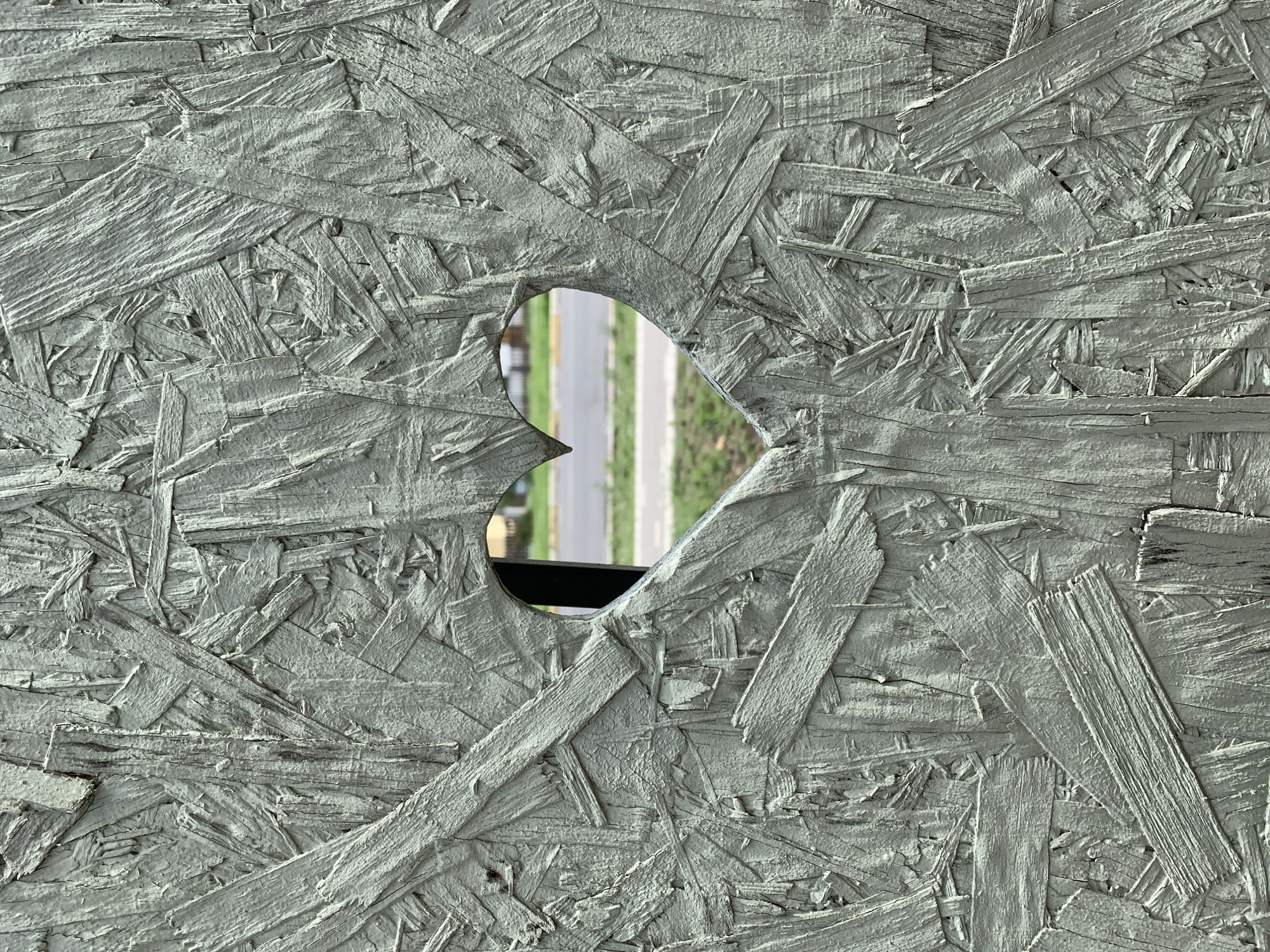 A heart-shaped opening in a light gray, rough-textured particle board wall reveals a blurred view of an urban environment. Beyond the opening, a paved road is visible, with a yellow line marking the center. Green grass and some indistinct structures are present in the background, suggesting an outdoor setting in a town or city. The particle board is composed of tightly packed, irregularly shaped wood chips, giving the surface a highly textured and weathered appearance. The scene appears to be in Hostomel, Ukraine.