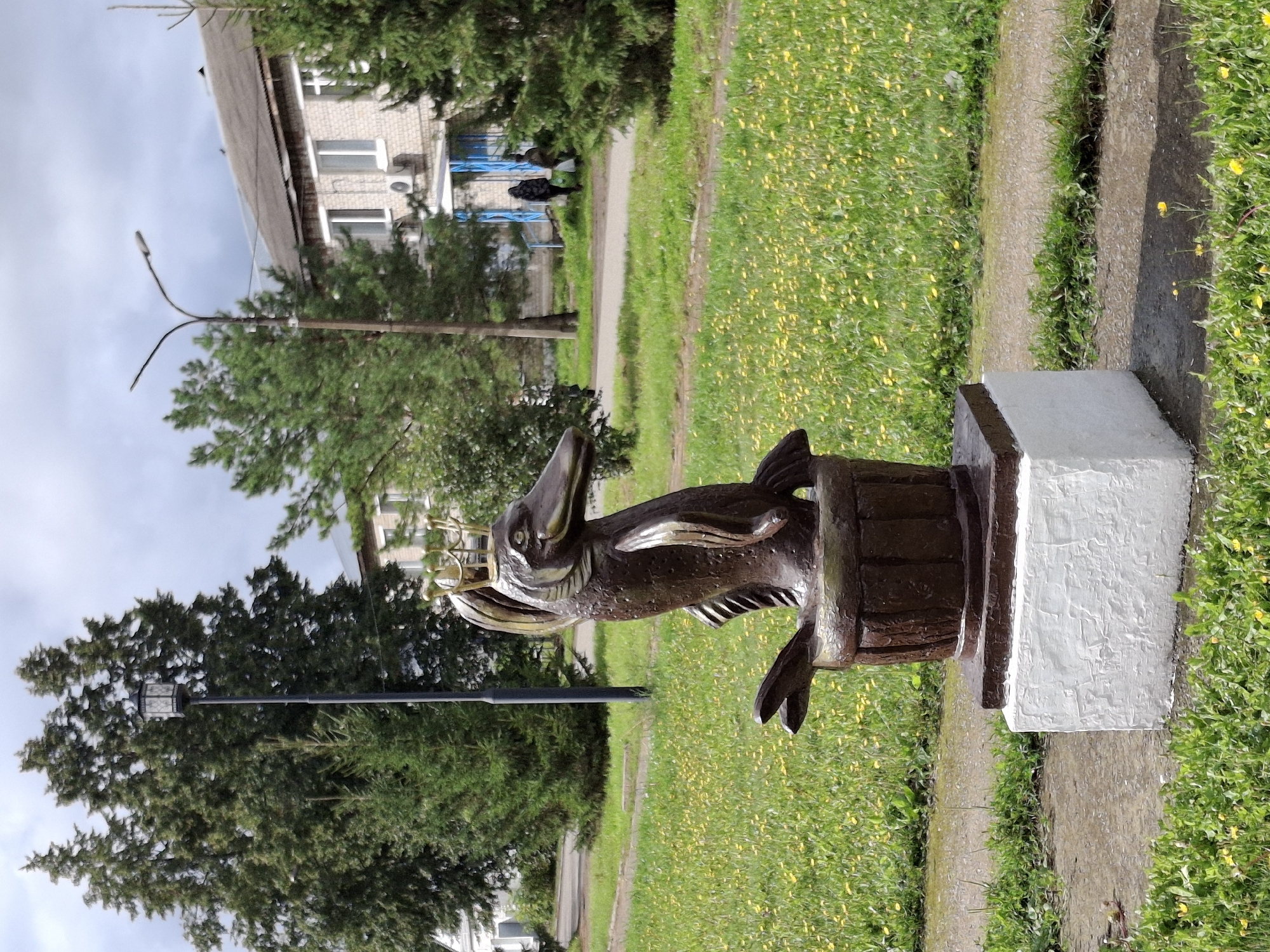 The image shows a bronze sculpture of a mythical creature, possibly a mermaid or a creature with fish-like attributes, wearing a crown, standing in a wooden barrel. The sculpture is mounted on a white rectangular pedestal. It is situated in a park-like area with a grassy lawn dotted with yellow flowers. In the background, there are trees, a lamppost, and a two-story brick building with multiple windows. Two individuals are visible in the distance walking on a path near the building. The setting appears to be a public space in the village of Elovo, Russia, based on the provided context.