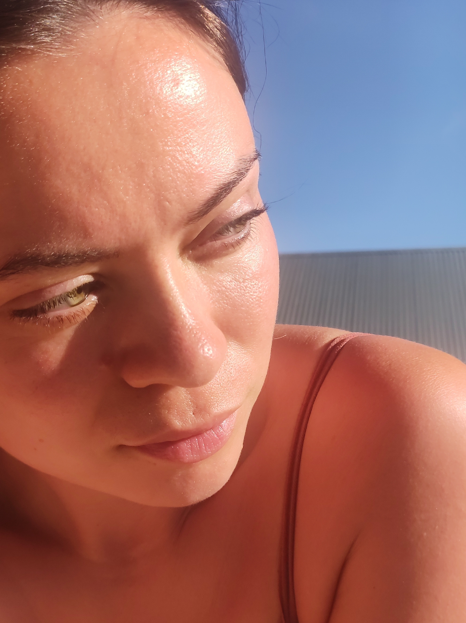A close-up photograph shows a woman with fair skin, green eyes, and dark hair pulled back. The sun is shining brightly, casting a warm glow on her face and shoulder, and there is a building with corrugated metal siding in the background to the right. The sky behind the building is a clear blue. She is wearing a thin-strapped top.