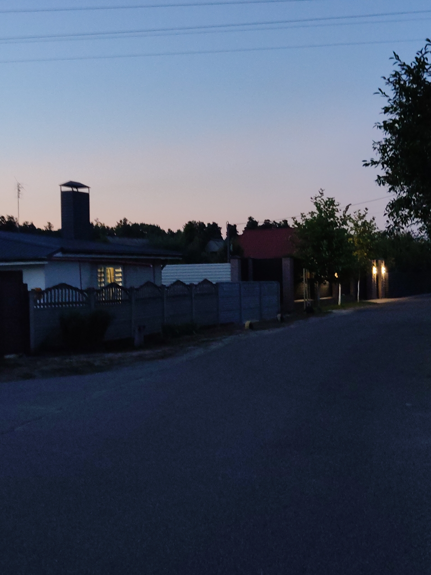 This image depicts a suburban street scene at dusk in Cherkasy, Ukraine. The sky is a gradient of pale blue to soft pink, with power lines visible near the top. On the left, a low-slung house with a dark roof and a prominent chimney is visible. One window of the house is illuminated from within. A grey, segmented fence runs along the front of the property. Trees and a darker building with a red roof are situated behind this house. Further down the street, on the right side, two illuminated outdoor lights are visible, casting a glow on the surrounding trees and a dark fence. The foreground is dominated by a dark asphalt road. The overall atmosphere is quiet and tranquil, characteristic of a residential area at the end of the day.