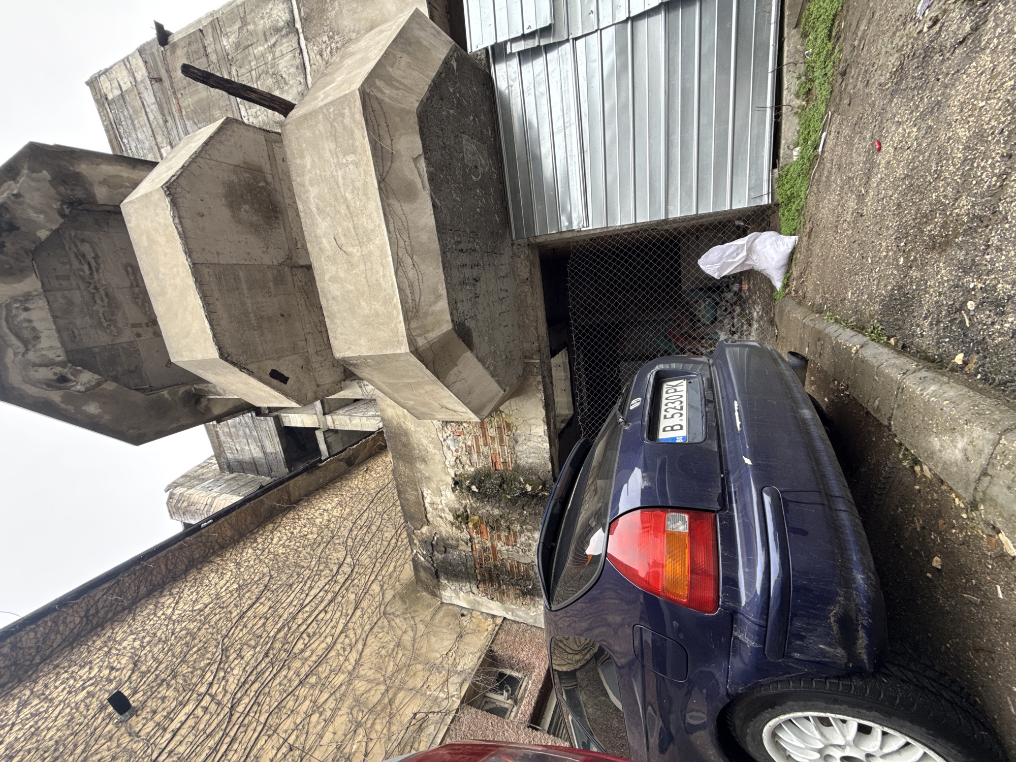 A low-angle shot captures a gritty urban scene in Shumen, Bulgaria. A hulking, layered concrete structure dominates the left side of the frame, displaying a brutalist aesthetic with its repeating geometric shapes and worn surfaces. An adjacent wall, partially covered in dried vines, adds to the aged ambiance. To the right of the concrete structure, a galvanized metal wall stands, fronting a chain-link fence. A dark blue car, visibly scuffed and marked, is parked closely against the curb, its rear section occupying the center-right. The license plate, bearing the registration "B 5230 PK", is clearly visible. A white plastic bag lies discarded near the base of the metal wall. The pavement, marked with stains and moss, suggests a damp, unkempt environment. The composition emphasizes the utilitarian and somewhat neglected aspect of this location in Shumen.