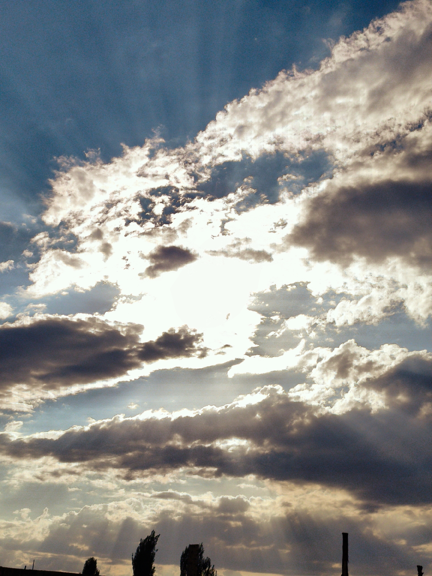 The image shows a dramatic sky filled with various types of clouds, illuminated by bright sunlight. Rays of light pierce through breaks in the clouds, creating a visual effect of light shafts. The lower portion of the image reveals a dark silhouette of a cityscape, with what appear to be trees or industrial structures along the horizon. One distinct, dark vertical structure, possibly a chimney or pillar, is visible on the right side. The overall impression is of a dynamic and somewhat moody sky above an urban or industrial landscape.