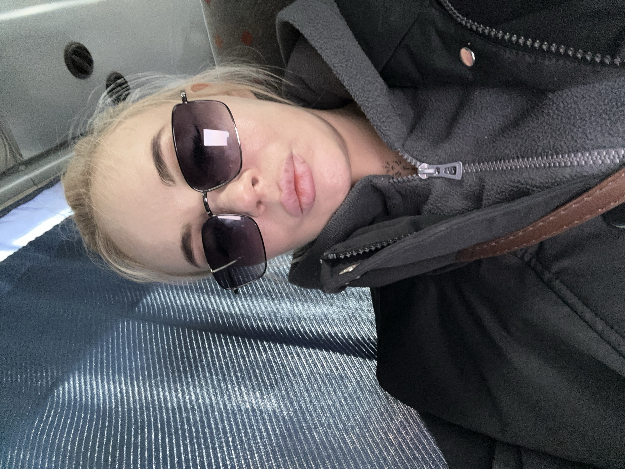 A blond woman wearing sunglasses and a black hooded jacket sits on a blue, striped seat. The location is unspecified, although the prompt suggests Megion, Russia. The woman's lips are pursed, and a tattoo is visible on her neck. The jacket is zipped partially open, revealing a gray fleece lining. A brown strap is also visible.