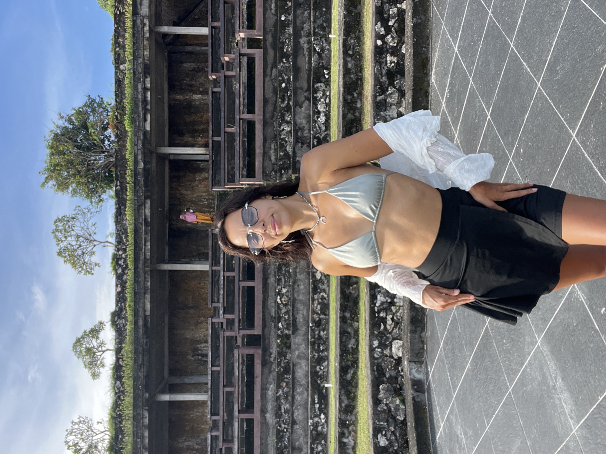 A young woman is captured in a bright, outdoor setting, posing slightly off-center to the right of the frame, smiling subtly towards the viewer. She wears a light sage green bikini top, dark black shorts or a skirt, and a sheer white long-sleeved cover-up. Her accessories include dark rectangular sunglasses, a silver necklace with a crescent moon pendant, and small earrings. Her right hand rests on her hip, while her left hand gently holds the fabric of her cover-up. Her dark hair is casually styled.
The background features a large, ancient-looking stone structure, possibly a temple or terraced ruins, characterized by multiple layers and steps made of dark, weathered stone. Green moss and small trees grow on and around the structure, indicating age and natural integration. The sky above is a clear, bright blue with minimal cloud cover, suggesting a sunny day. The foreground consists of a dark grey tiled surface with white diagonal grid lines, serving as the ground she stands on. The bright lighting indicates it is likely midday or early afternoon. The scene conveys a relaxed, summery atmosphere, typical of outdoor exploration in a warm climate like Kuta Selatan, Indonesia. No discernible text is visible in the image.