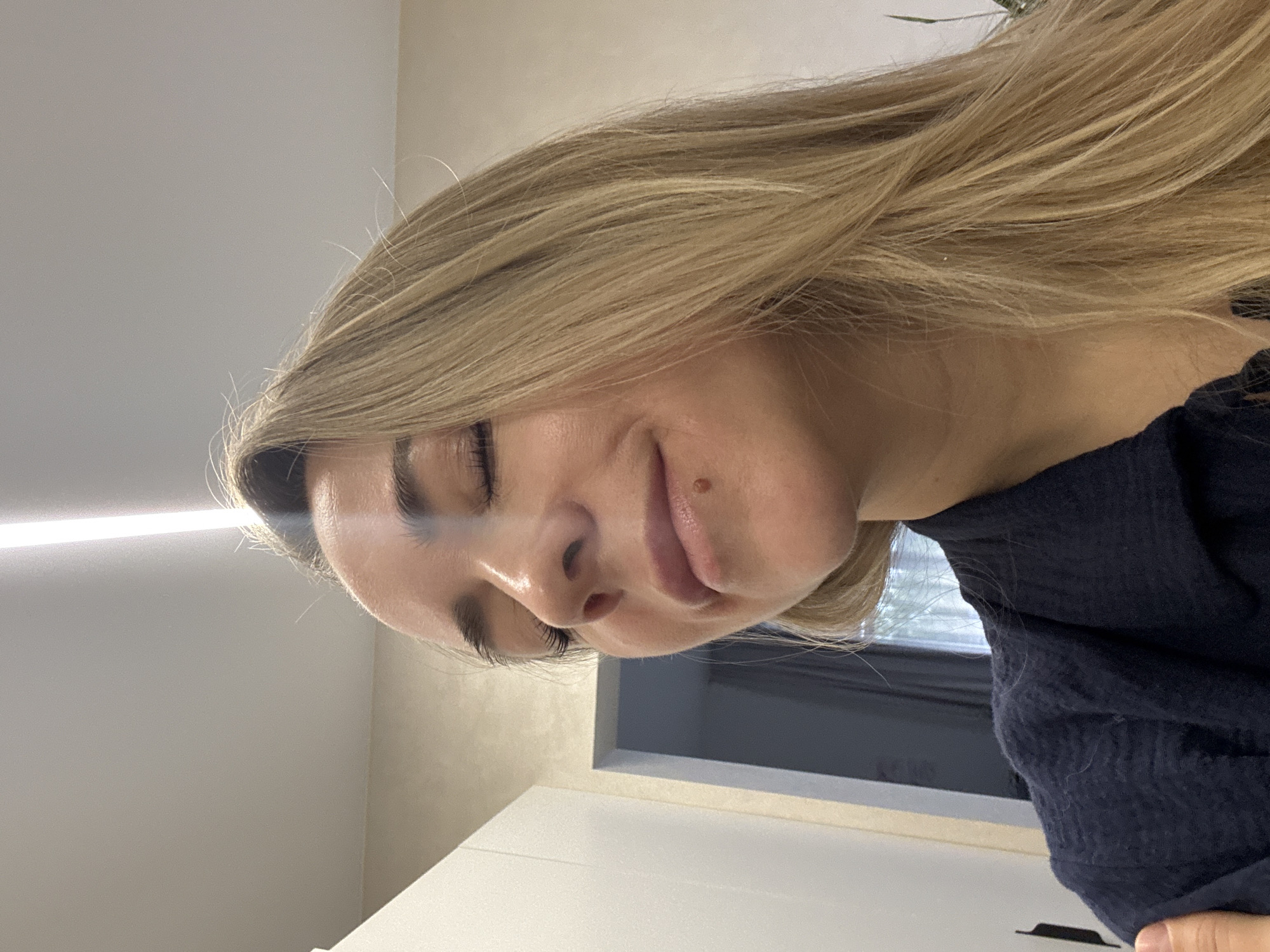 The media file displays a headshot of a woman, rotated 90 degrees clockwise. The woman has long, straight or slightly wavy blonde hair with lighter highlights, which cascades around her. She has fair skin, with her eyes appearing closed or cast downwards, and dark, well-defined eyebrows and eyelashes. A small, dark mole or beauty mark is visible above her upper lip, slightly to the right side of her face. Her lips are softly closed, and a gentle, serene smile plays on her face, conveying a sense of calm or relaxation.
She is wearing a dark blue or black textured garment, possibly a sweater, visible around her neck and shoulder area. The background suggests a modern indoor setting. A plain, light-colored wall is visible behind her. To the left side of the frame (when viewing the rotated image), a bright horizontal strip of light, likely an LED fixture, illuminates the scene. A dark window frame and a glimpse of what appears to be an outdoor view can be seen further back. Part of a white surface, possibly a cabinet or counter, is in the foreground. The lighting is bright and even, indicating daytime. There is no visible text in the image. The scene is located in Chaikovsky, Russia.