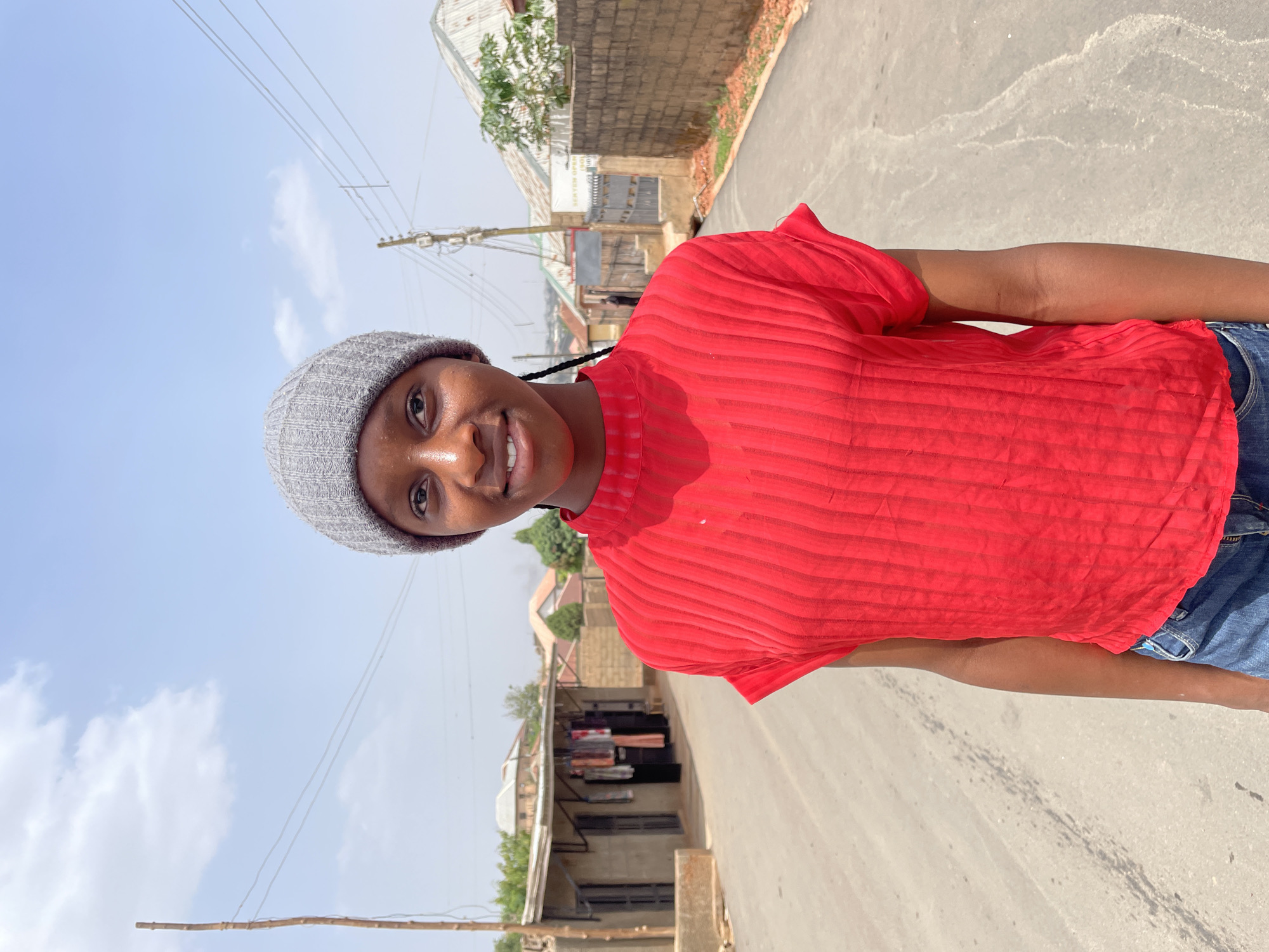 A young woman is standing on a paved road, facing the camera with a slight smile. She is wearing a gray beanie, a red horizontally striped short-sleeved shirt, and blue jeans. The road stretches behind her, leading to a blurred background of buildings and vegetation. Power lines are visible against a light blue sky. The location appears to be a street in Jos, Nigeria, based on the architectural style and surrounding environment.