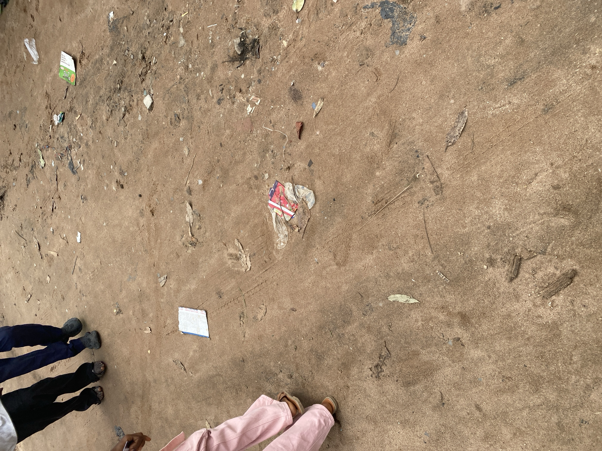 The image shows a ground covered in dirt and debris, with several people's legs and feet visible. The people are wearing dark pants and shoes, and one person is wearing pink pants and sandals. They appear to be standing on a dirt or gravel surface, possibly outdoors. There is scattered trash on the ground, including a crumpled piece of paper with some text that appears to be in English. The lighting suggests it is daytime. The scene is likely a public space in Jalingo, Nigeria, given the location context. No specific activity or event is clearly depicted, beyond people standing around.