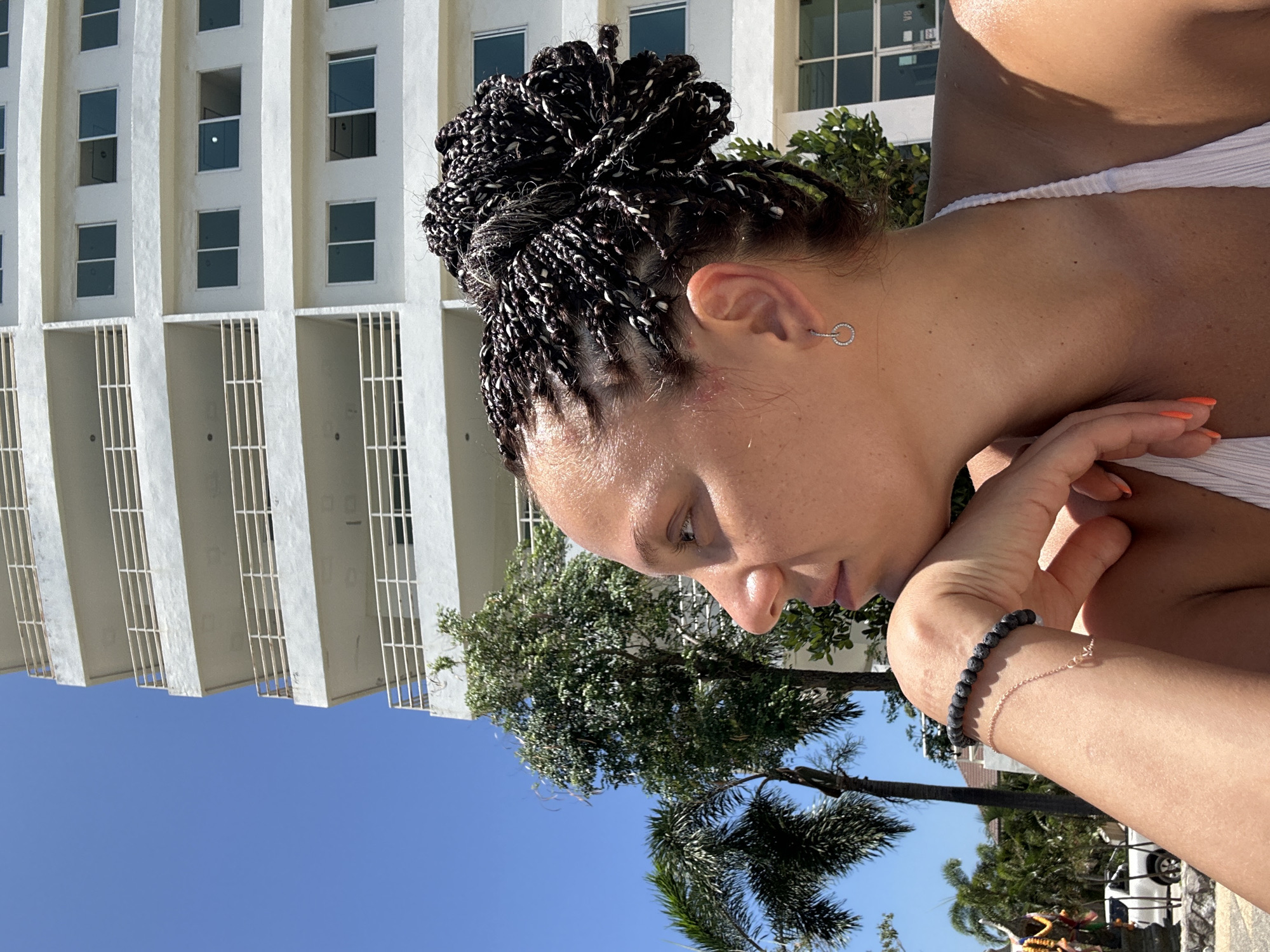 The image presents a close-up, profile view of a person, likely female, captured outdoors under bright sunlight. The person has a dark skin tone and their head is tilted downwards, suggesting a contemplative or relaxed moment. Their hair is styled in numerous dark braided twists, some with white strands, gathered into a bun on top of their head. They wear a simple silver hoop earring in their right ear and a white ribbed top or swimsuit. On their right wrist, two bracelets are visible: one made of dark, textured beads, and a delicate rose gold-toned chain. The fingernails on their right hand are painted a vibrant orange.
The background features a tall, modern white building with rows of dark-paned windows and balcony structures with vertical white railings. Lush green foliage, including tropical palm trees, is prominently displayed between the subject and the building, and also towards the lower right of the frame. The sky above is clear and bright blue, indicative of a sunny day. A white vehicle and a colorful, indistinct object are partially visible in the far background on the right. The overall setting is consistent with a tropical location like Pattaya City, Thailand. There is no visible text in the image.
