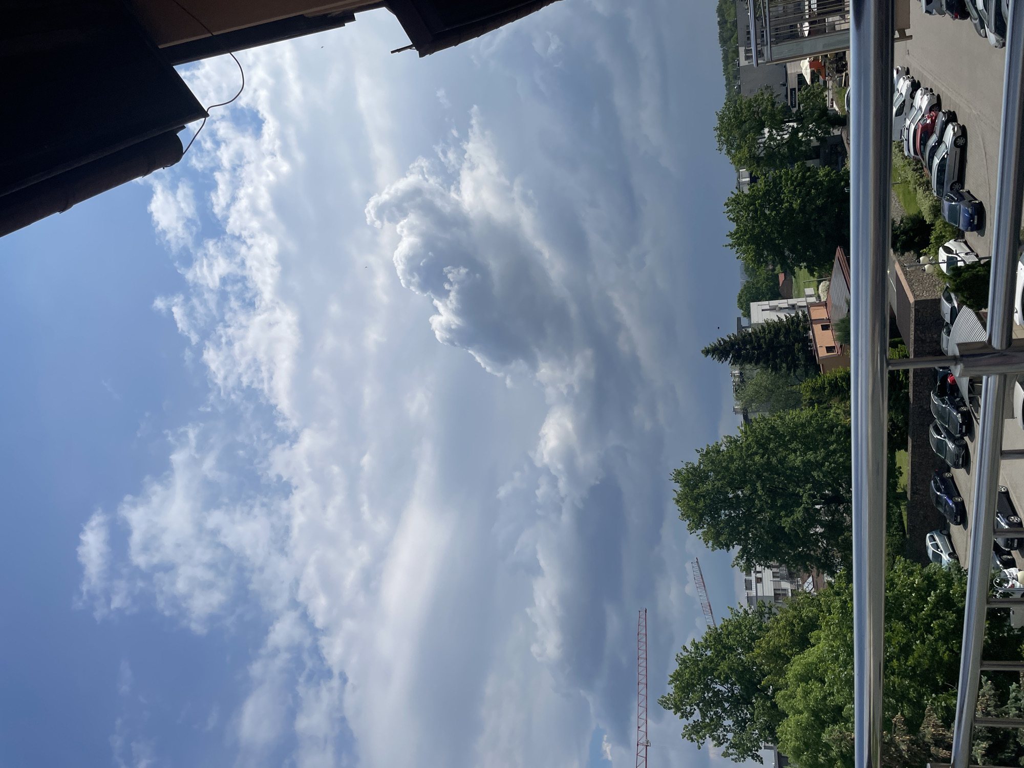 View of a sky with a mix of cumulus and stratus clouds, partially obscured by an overhanging roof edge in the upper left corner. Trees and buildings are visible on the right side of the image, along with a parking lot containing several cars. Construction cranes rise above the treeline. A balcony railing is present in the foreground. The scene appears to be in an urban environment.