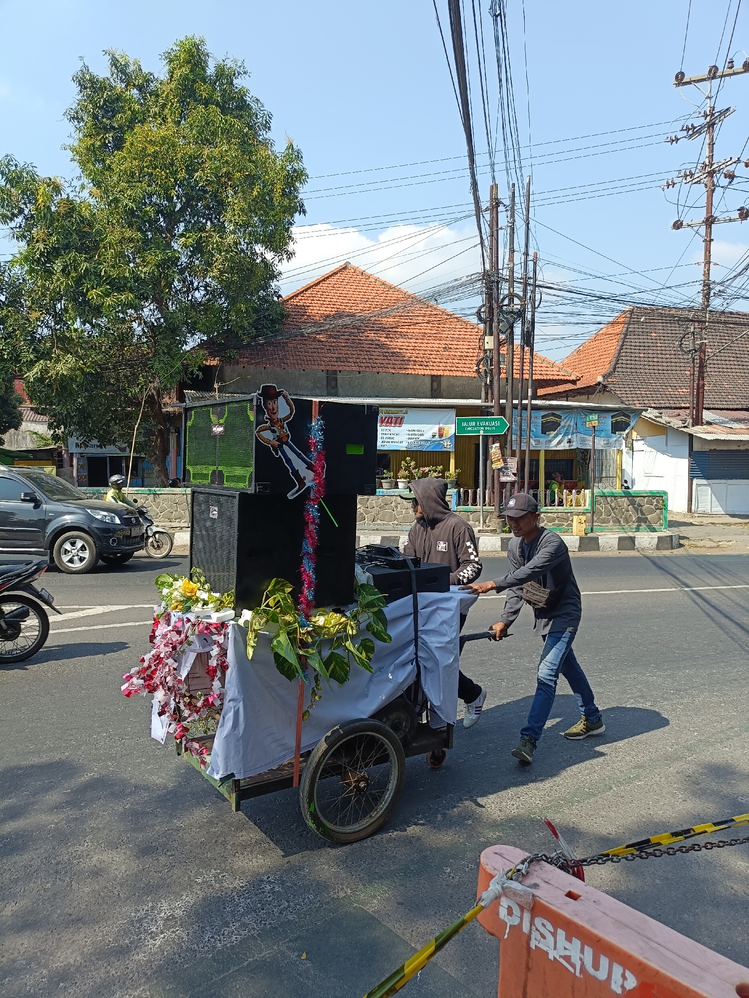 In the city of Kediri, Indonesia, two individuals are seen pushing a cart laden with audio equipment. The cart features two large speakers, one of which is adorned with a cutout image of Woody from Toy Story, and is decorated with garlands of red, white, and blue tinsel. Additional floral arrangements and greenery are placed on the cart. The cart is covered with a light-colored cloth. One person, wearing a dark hoodie with white checkered sleeves and a black baseball cap, is positioned on the left side of the cart, while the other, in a grey long-sleeved shirt, blue jeans, and a black baseball cap, is pushing the cart from the right. They are on a paved road, likely a street, with a black SUV and a motorcycle visible in the background. Further back, several buildings with red-tiled roofs and various signage are present, including what appears to be a shop. Overhead, a complex network of utility poles and wires stretches across the sky, indicating an urban or semi-urban environment. The ground displays tire tracks and what looks like a section of a construction barrier with yellow and black hazard tape. The scene is illuminated by bright daylight, suggesting a clear sky.