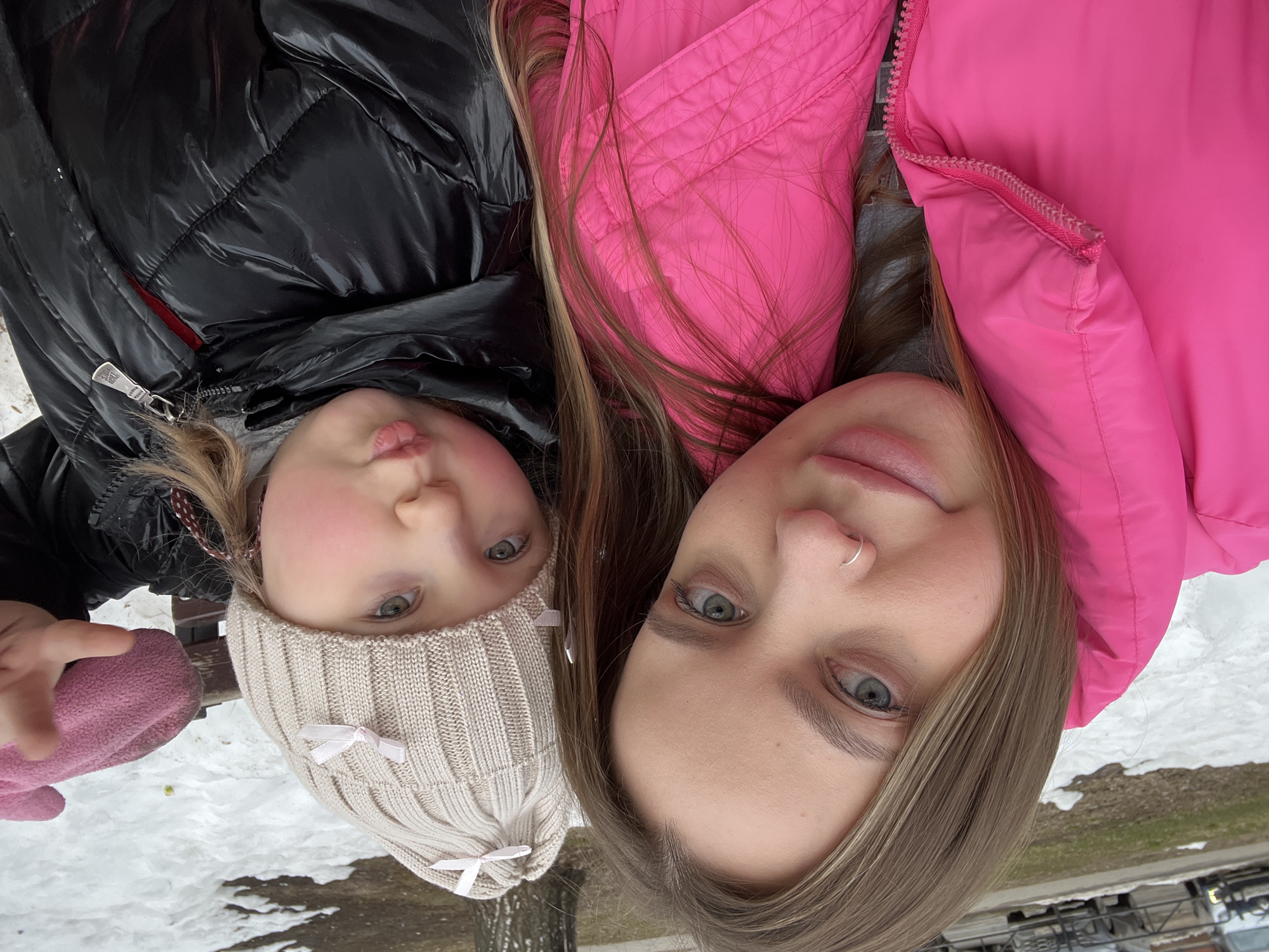 A close-up outdoor selfie depicts an adult female and a young child, likely in a park or recreational area within Chaikovsky, Russia, during a late winter or early spring season. The adult, positioned on the left, has long light brown hair and light eyes, wearing a bright pink puffer jacket and a silver nose ring on her left nostril. She looks directly at the camera with a subtle smile. The child, on the right, wears a cream-colored knit beanie adorned with multiple small pink ribbon bows and a shiny black puffer jacket with a visible red lining. The child's lips are pursed, and their right hand, covered by a pink glove, is raised and pointing towards the camera. The background reveals patchy, melting snow on the ground, a few bare trees, a dark wooden or metal bench, and indistinct parked vehicles in the far distance on a paved surface. A small yellow object is visible on the snow.