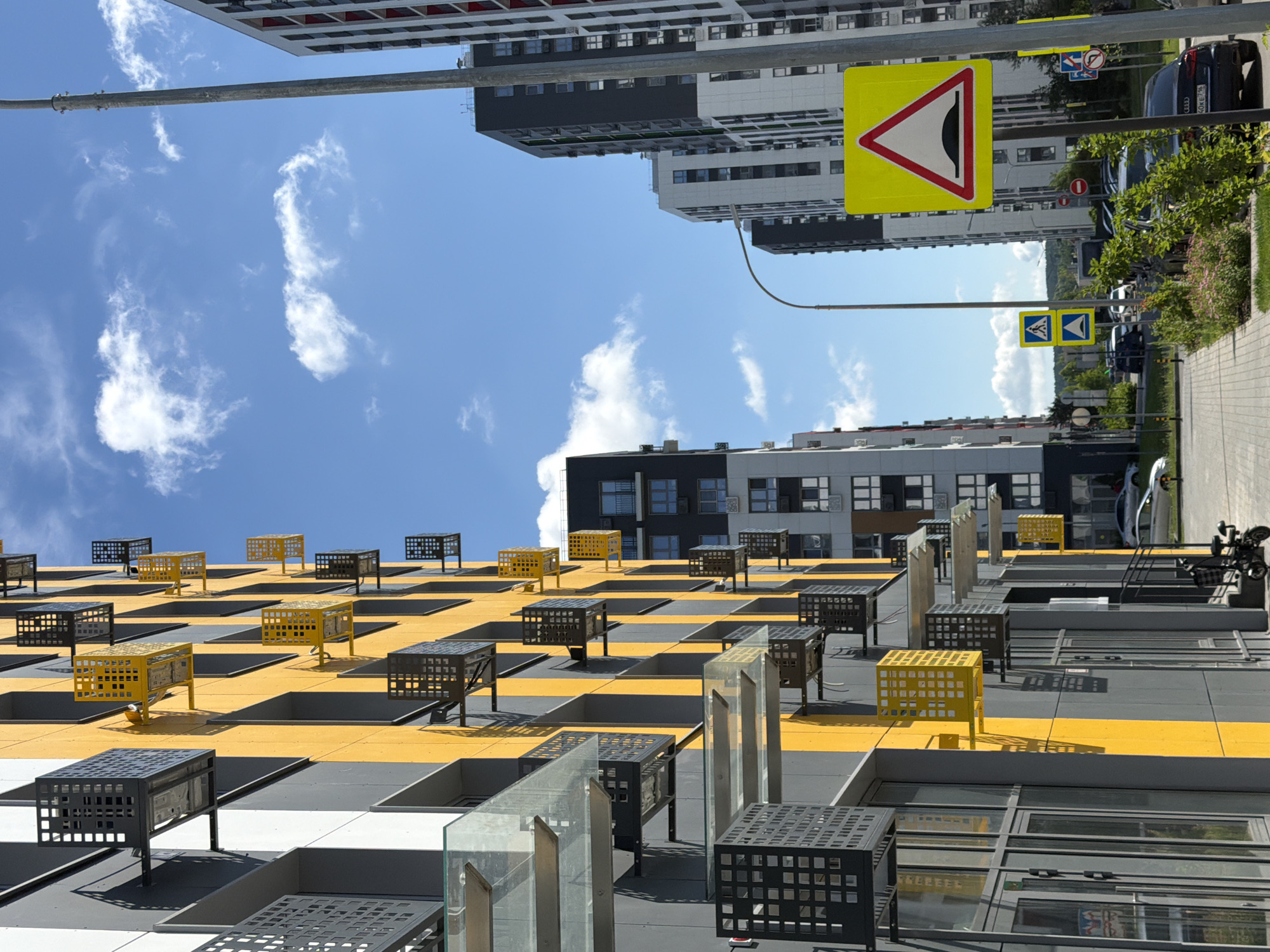 In an outdoor urban environment, a modern apartment building with a distinctive yellow and grey facade dominates the left side of the frame. The building features numerous balconies with black and yellow metal enclosures. Tall apartment buildings with dark grey and white exteriors rise in the background, creating a dense urban landscape under a bright blue sky with scattered white clouds.
In the foreground, a paved walkway lines the street, bordered by lush green bushes and flowering plants. Several cars are parked along the street, including a black Audi sedan. Traffic signs are visible, including a yellow triangular sign with a red border indicating a speed bump. A lamppost stands to the right of the frame. The scene is under bright daylight.
