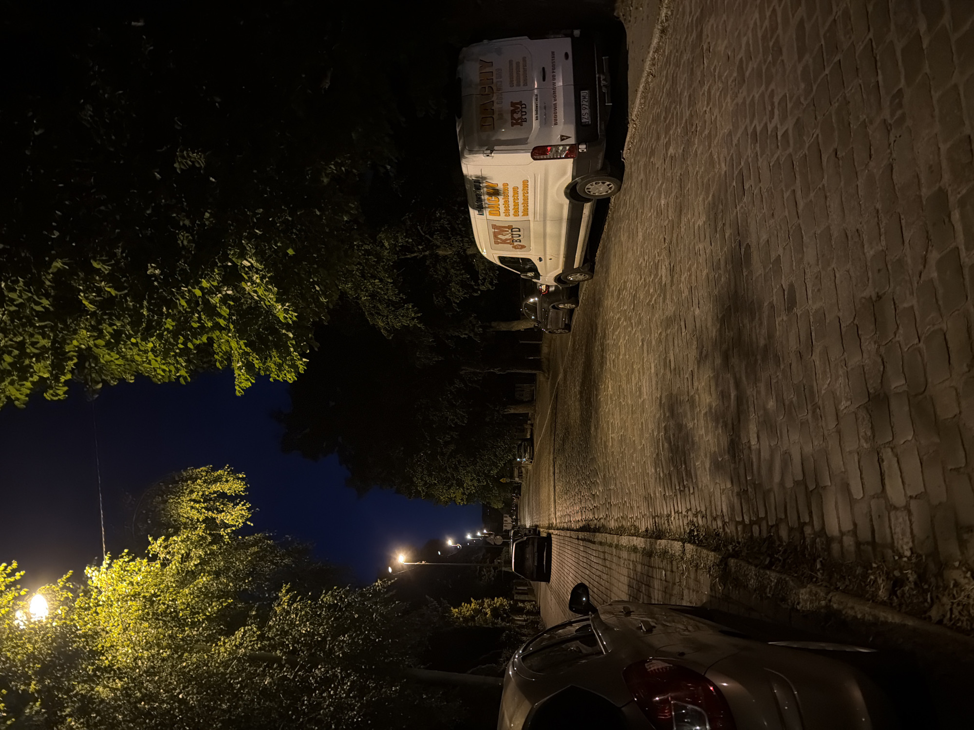 The image depicts a street scene at night in Szczecin, Poland, likely in a residential area. A cobblestone street runs diagonally from the foreground to the background, illuminated by streetlights. On the left, a silver car is parked on the side of the road. Further down the street, several other cars are parked along the sidewalk, which is also made of cobblestones. On the right side of the street, a white van with logos and text in Polish is parked. The van's signage suggests it is related to construction services, specifically mentioning "DACHY" (roofs) and "KM BUD." Trees line both sides of the street, their leaves illuminated by the streetlights, casting shadows on the road. The sky is dark blue, indicating nighttime. The overall atmosphere is quiet and still.