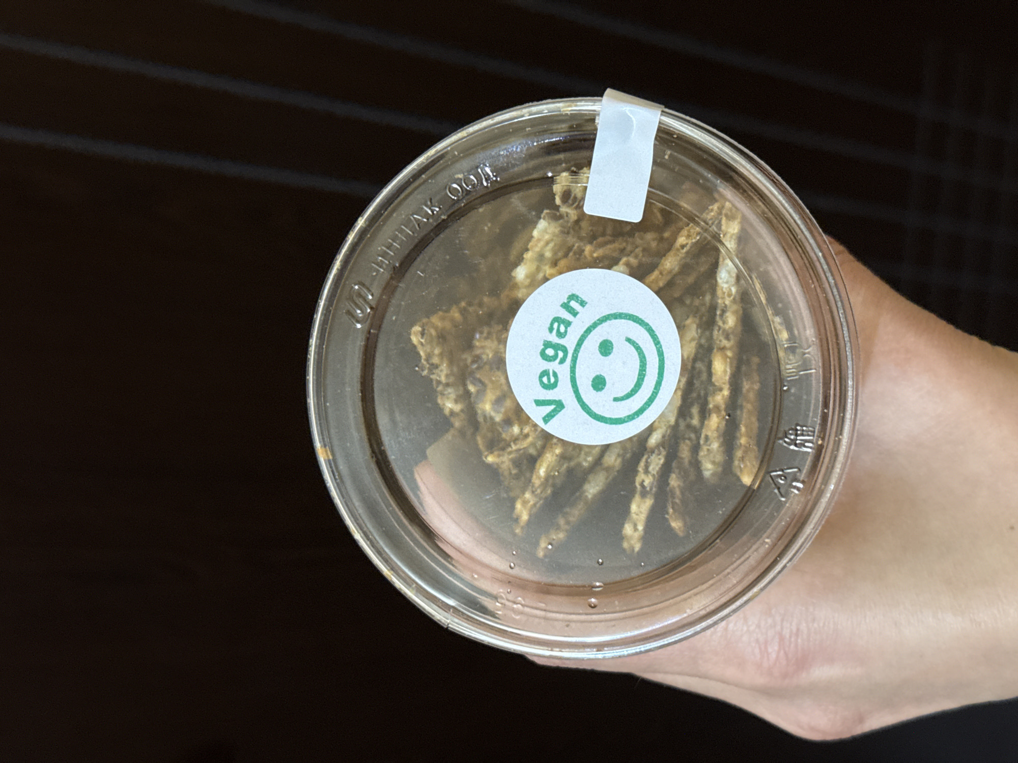 A person is holding a clear, round, plastic container filled with brown, stick-like food items. A circular, green and white "Vegan" sticker with a smiley face is affixed to the lid. A small, white sticker is also adhered to the lid near the container's edge. The container is being held against a dark brown, possibly wooden surface. The photo appears to be taken indoors. There is no evidence in the image to suggest the location is in the city of Pleven, Bulgaria.